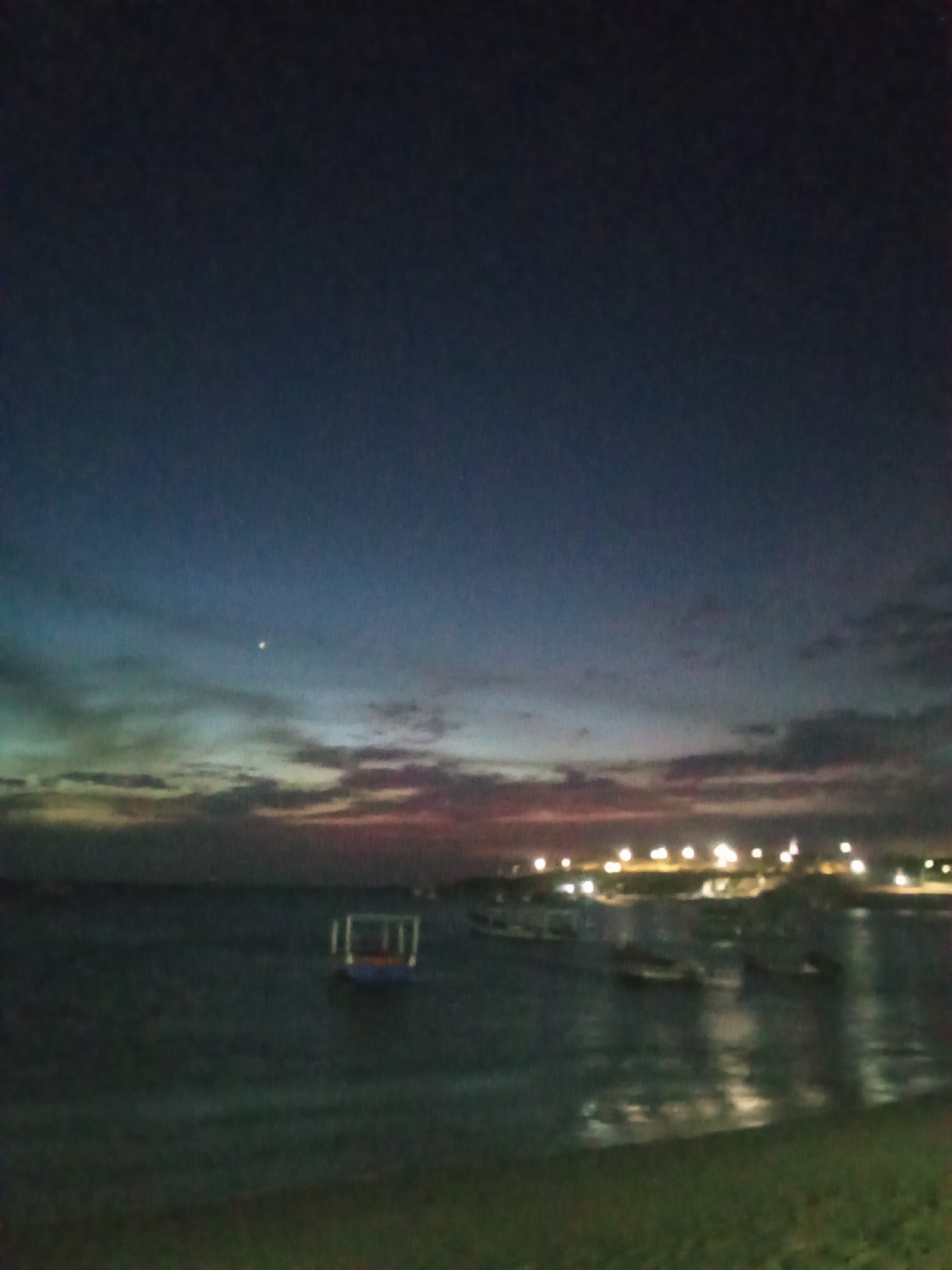 The image depicts a nighttime view of the coastline at Juan Griego, Venezuela. The sky is a deep blue, transitioning to lighter hues of pink and green near the horizon, where clouds are present. A small, faint object, possibly the moon or a star, is visible in the sky. The water is dark and reflects the light from the shoreline. Several boats are scattered in the water. Lights are visible along the coastline, suggesting buildings or infrastructure. The foreground shows a grassy area.