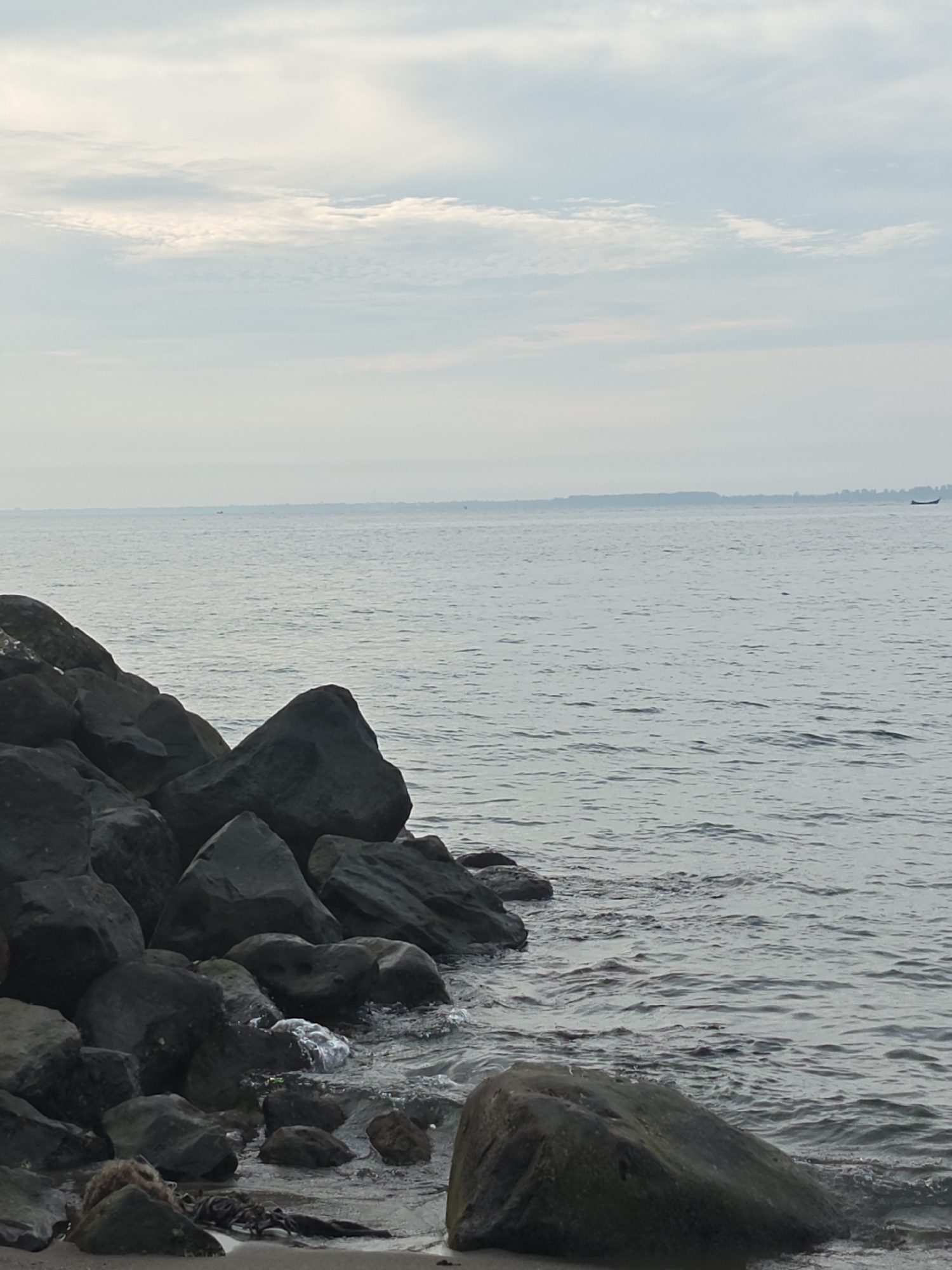 The image shows a rocky shoreline on a cloudy day, with a calm body of water stretching to the horizon. The rocks are dark and varied in size, with some closer to the camera and others forming a breakwater further out. The water is a muted gray, with gentle ripples. In the distance, a faint line of trees or land is visible, and a small boat can be seen on the water. The sky is filled with soft, layered clouds in shades of gray and white. The scene appears to be a natural landscape, likely a beach or coastal area. This location could be the shore of the Malacca Strait in Lhokseumawe, Indonesia, known for its natural beauty and coastal features. There are no people visible in the image.