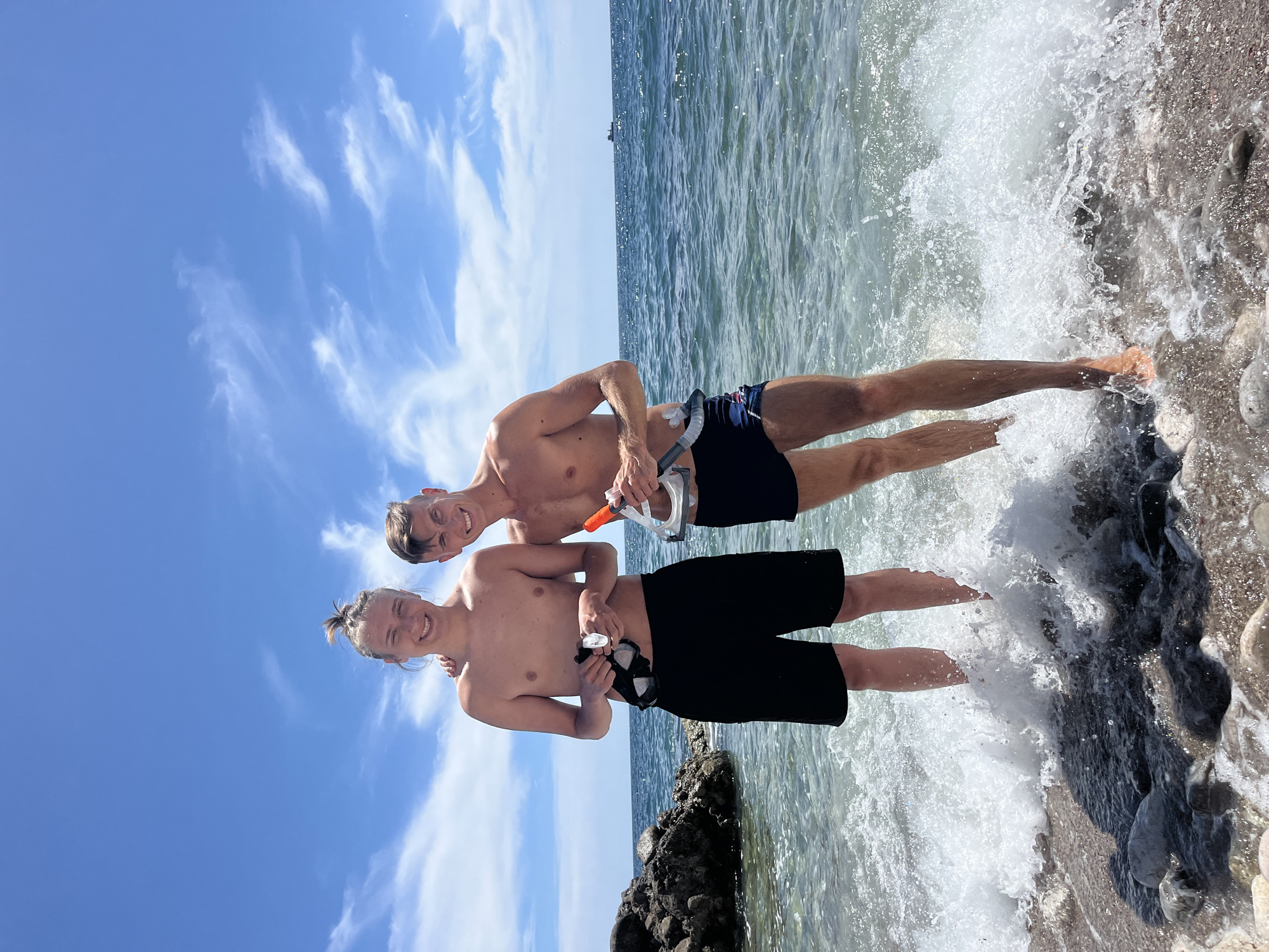 Two young men stand knee-deep in the sea at a pebble beach, with waves breaking around them. They are both shirtless and wearing swim trunks, and each holds a snorkeling mask and snorkel. The man on the left has light brown hair pulled up in a bun and wears black swim trunks. The man on the right has shorter, darker hair and wears navy blue swim trunks with a red and white stripe. Behind them, the calm sea extends to the horizon under a bright blue sky with scattered white clouds. In the distance, a cargo ship is visible. To the left of the frame, large rocks are visible at the water's edge. The scene appears to be a sunny day at a beach in Bar, Montenegro.