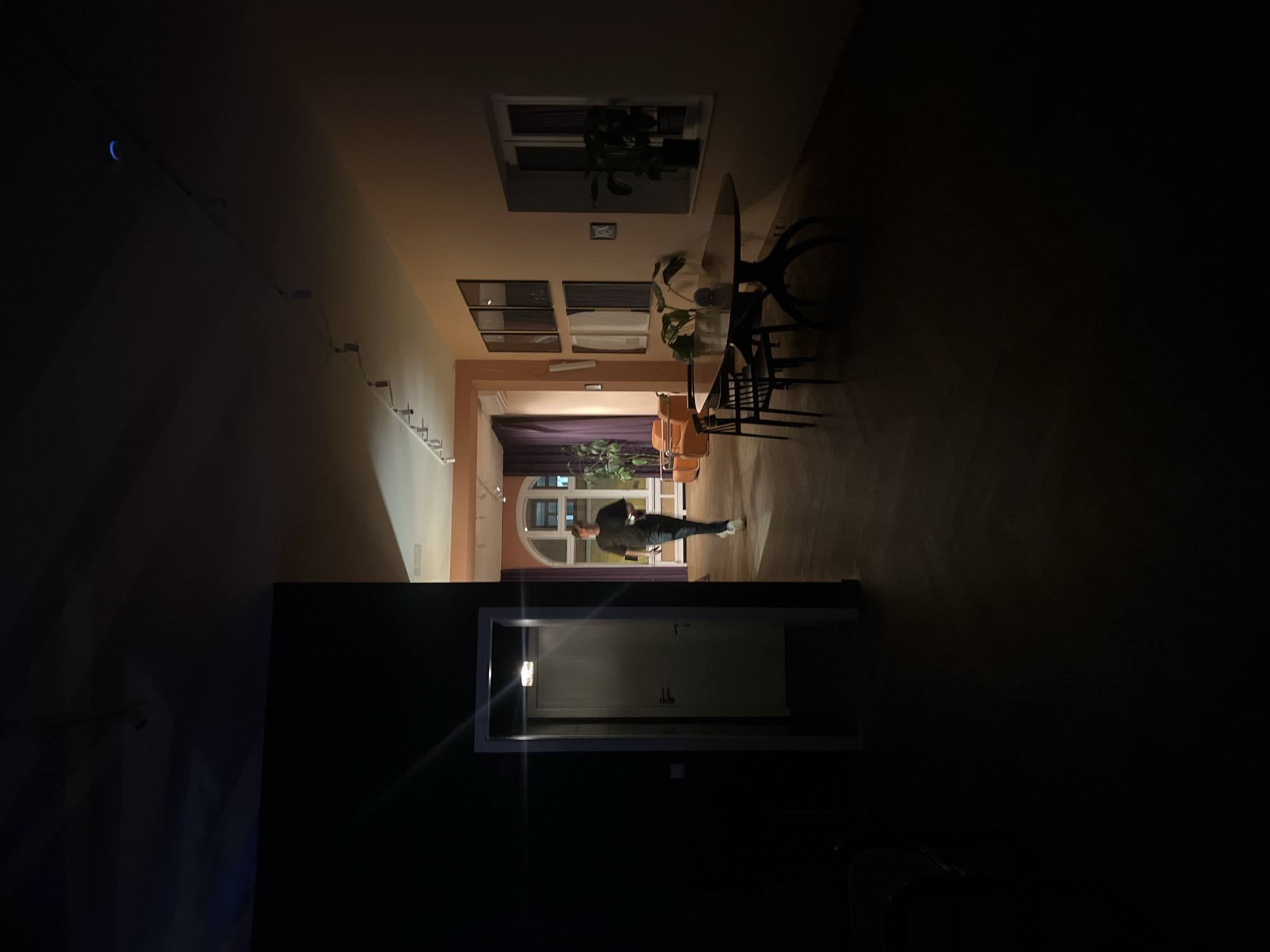 The image displays an interior view of a building in Berlin, Germany, under low light conditions. The environment features dark herringbone-patterned wooden flooring and walls painted in a light peach or orange tone in the illuminated sections.
In the foreground, a brightly framed white door is open on the left, revealing a dimly lit interior. A light fixture is visible above this doorway, emitting a star-shaped lens flare.
A male individual, appearing to be in his 20s or 30s, is captured in motion, walking from left to right in the mid-ground. He is dressed in a dark t-shirt, dark trousers, and white athletic shoes, and holds a small white object in his left hand. His face is discernible but slightly blurred.
To the right, a dining area is visible, furnished with an oval-shaped wooden table featuring a dark, sculptural base, and two dark wooden chairs. A potted plant is situated on the table. The right wall contains a window with a potted plant on its sill, and three framed rectangular objects are mounted vertically. The ceiling features a track lighting system with multiple spotlights.
In the background, a large arched window or doorway is framed by deep purple curtains on either side. To the left of this archway, a lounge area includes two orange armchairs and a small table between them, accompanied by a tall potted plant. A small blue crescent light source is visible on the ceiling towards the upper right.