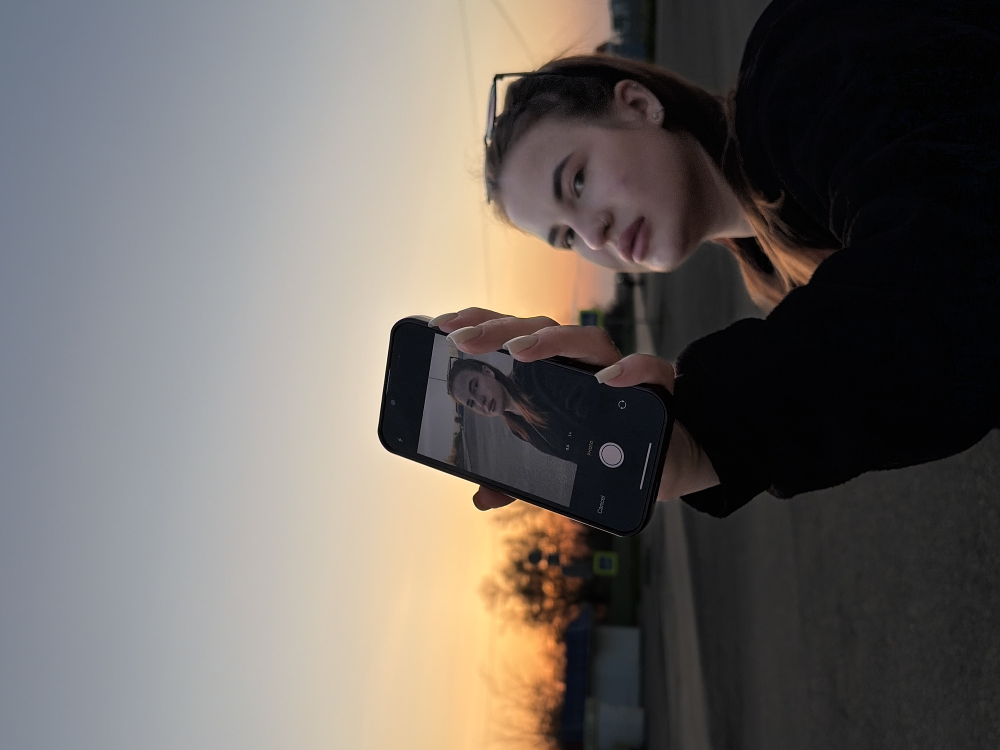 The image is a vertical shot featuring a young female taking a selfie or examining her reflection in a smartphone camera app, set outdoors in Novopokrovskaya, Russia. The scene is illuminated by the warm, soft light of either sunset or sunrise, casting a golden glow across the horizon.
The subject is a young woman with long brown hair, possibly partially tied back, and glasses resting on top of her head. She is wearing a dark top or jacket. Her expression is neutral and focused on the phone screen, held by her hands which display manicured nails.
She holds a dark-colored smartphone, likely an iPhone due to the visible interface elements. The phone screen clearly shows a camera app in selfie mode, displaying her own reflection. Visible text on the screen includes "0.5x", "1x", "PHOTO", a white circular shutter button, and a "Cancel" button. A small circular arrow icon for switching cameras is also present.
The background consists of a sky that transitions from a light blue-grey on the left to a warm, peachy-orange on the right, indicating the low angle of the sun. Below the horizon, the details are blurry, showing dark, indistinct shapes that suggest trees or distant structures against the warm light, and what appears to be a road or open ground. The weather appears clear. The overall impression is calm, centered around the act of self-photography during the "golden hour."