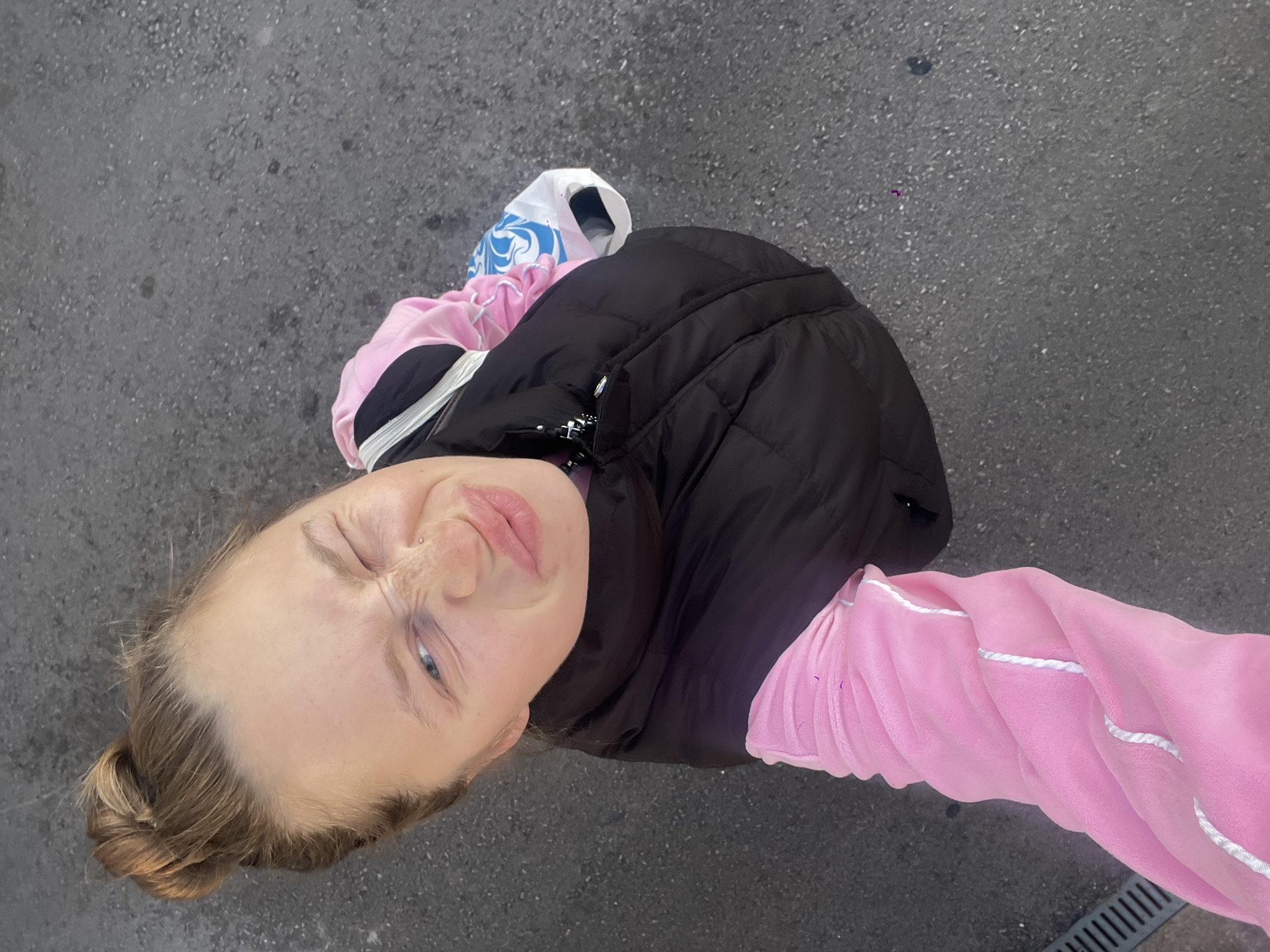 A selfie taken from an elevated angle shows a woman with blonde hair pulled back into a bun, wearing a black puffer vest over a pink sweater with white trim. She is standing on a paved surface, likely a sidewalk or street, holding shopping bags. The background suggests an urban environment in Bečići, Montenegro.