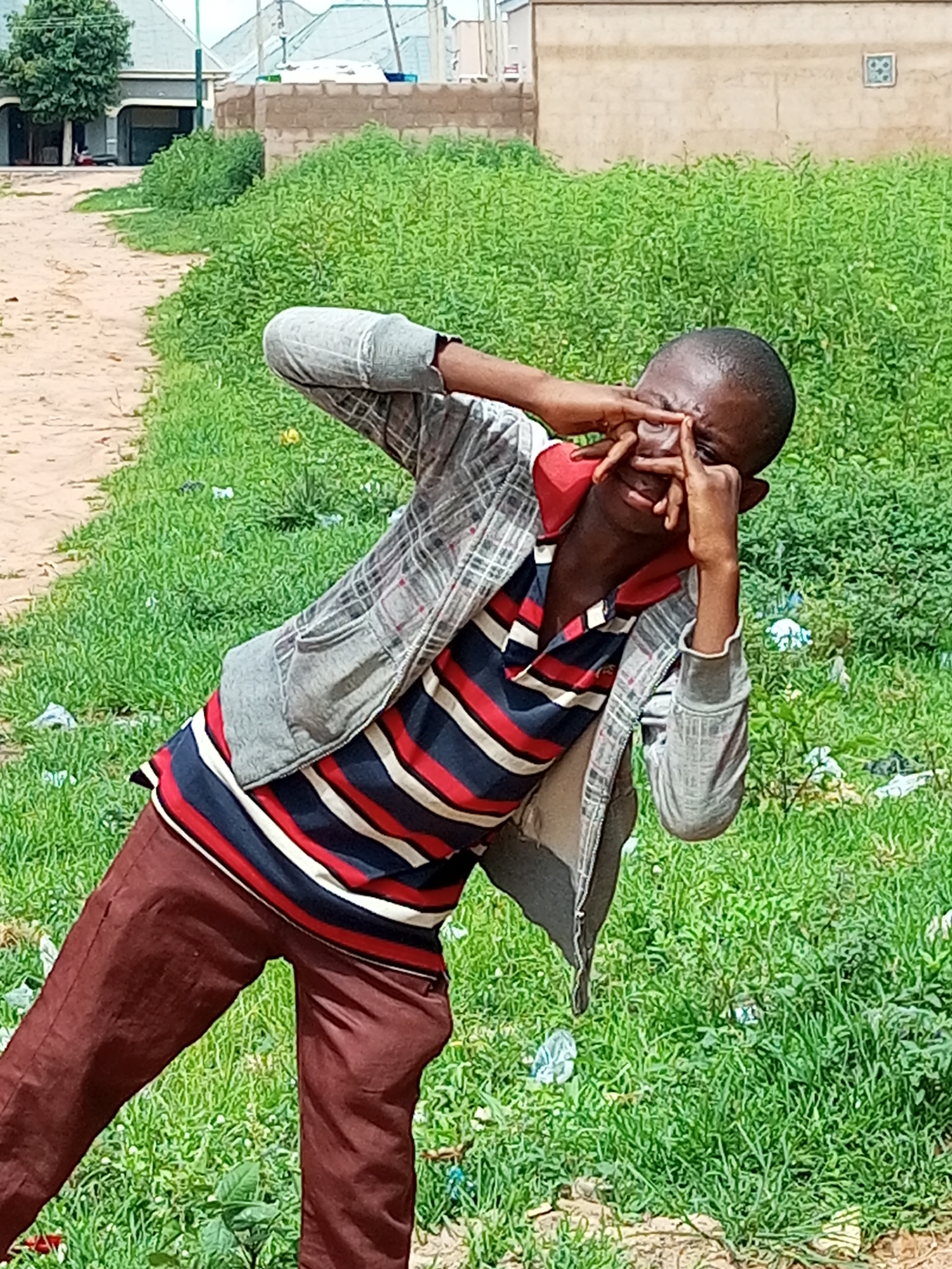 The image depicts a young Black male in Tempure, Nigeria, posing with his hands over his eyes, as if peeking. He is wearing a grey and red plaid jacket over a red, white, and blue striped polo shirt, and brown trousers. The background features a dirt path, lush green grass with some litter, and buildings with pitched roofs in the distance. The overall scene suggests an outdoor, possibly informal setting within a Nigerian urban or peri-urban environment.