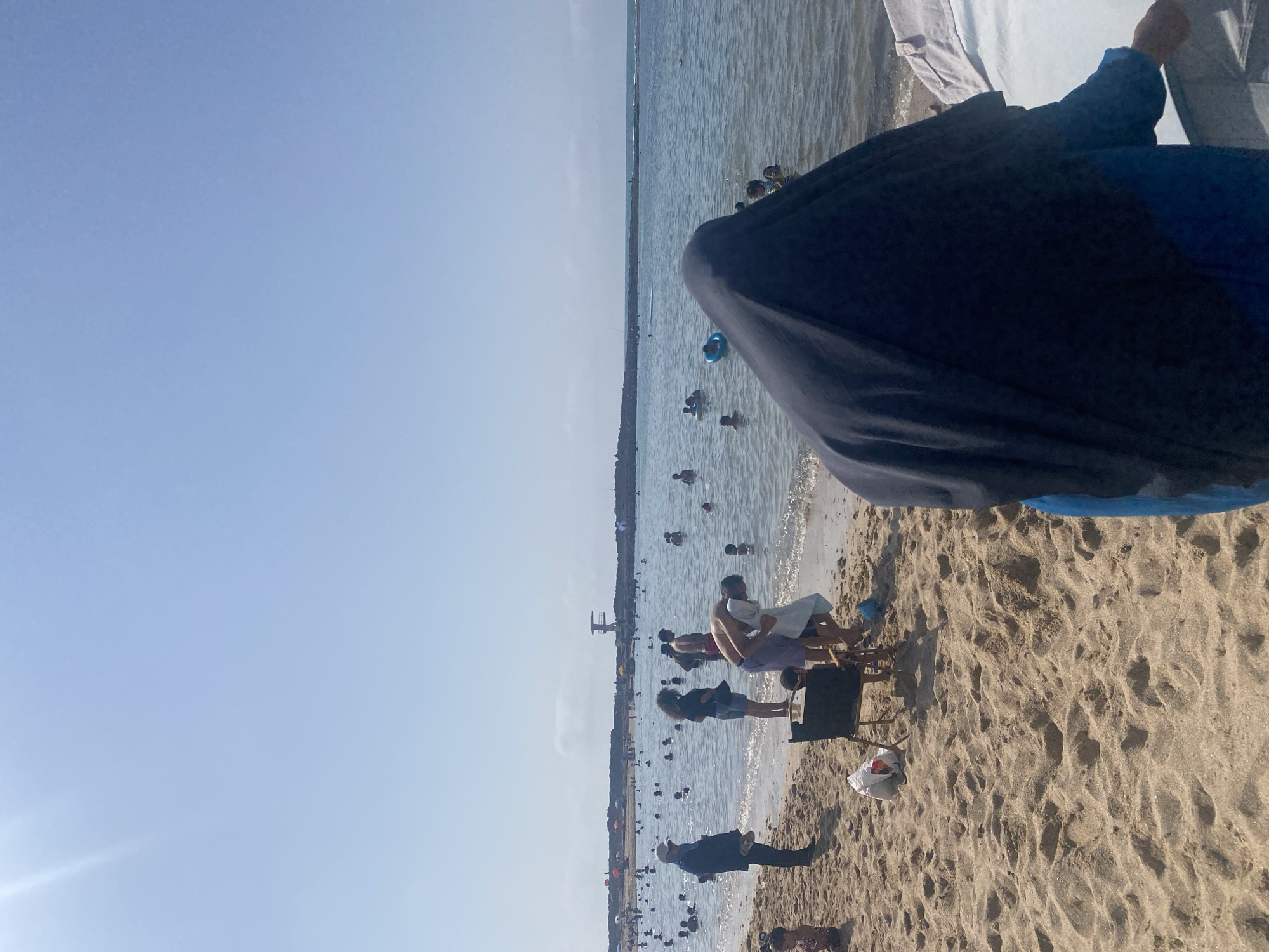 The image depicts a busy beach scene in Harhoura, Morocco, on a clear, sunny day. The foreground is dominated by sandy beach with footprints. In the middle ground, a diverse group of people are enjoying the water and beach activities. Several individuals are wading or swimming in the calm sea. Some are seen interacting with each other, while others are engaged in solo activities. Beach chairs and bags are scattered on the sand, indicating a leisurely day. In the background, a long stone jetty stretches into the sea, and a lifeguard tower is visible atop it, suggesting an organized and safe beach environment. The sky is a clear, pale blue, indicating good weather conditions. The overall impression is one of a lively and popular coastal resort.