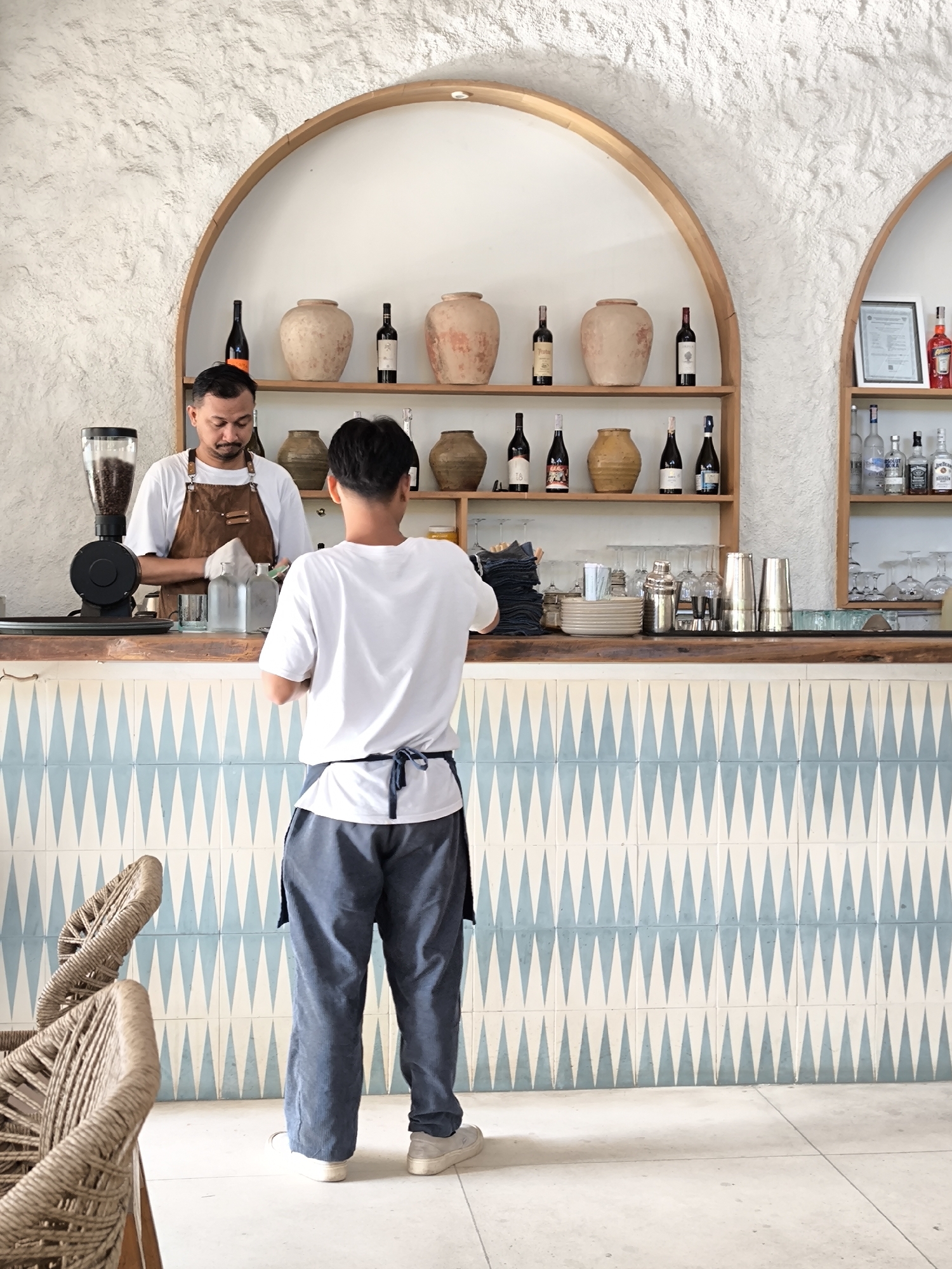 The photo shows the interior of a café or bar in Kute, Indonesia. Two men are visible. One is behind the counter, wearing a brown apron over a white t-shirt. He appears to be preparing drinks or food. The other man stands in front of the counter, facing the first. He is wearing a white t-shirt and blue trousers with a dark apron tied around his waist.
Behind the counter, there are shelves stocked with bottles, vases, and other decorative objects. A coffee grinder is visible on the left side of the counter. The front of the counter is covered with blue and white triangular patterned tiles. In the foreground, a portion of a wicker chair is visible. The walls are textured and white. The lighting appears to be natural.