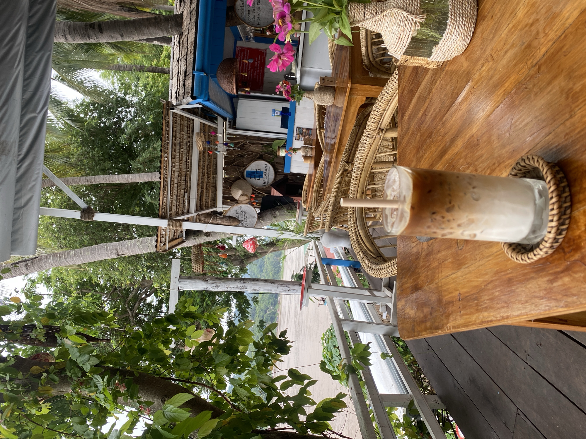 The image depicts a cafe with a rustic, tropical ambiance, likely located in Luang Prabang, Laos, overlooking the Mekong River. In the foreground, a wooden table holds a tall glass of iced coffee with layers of milk and espresso. The table is situated on a wooden deck or balcony, enclosed by a white railing. Beyond the table, wicker chairs surround more wooden tables, and further back, a counter area with a blue trim is visible, adorned with woven decorations and hanging tassels. The background reveals lush green foliage, palm trees, and the wide, sandy-colored Mekong River with distant hills. The overall scene suggests a relaxed outdoor dining or drinking experience in a natural setting.