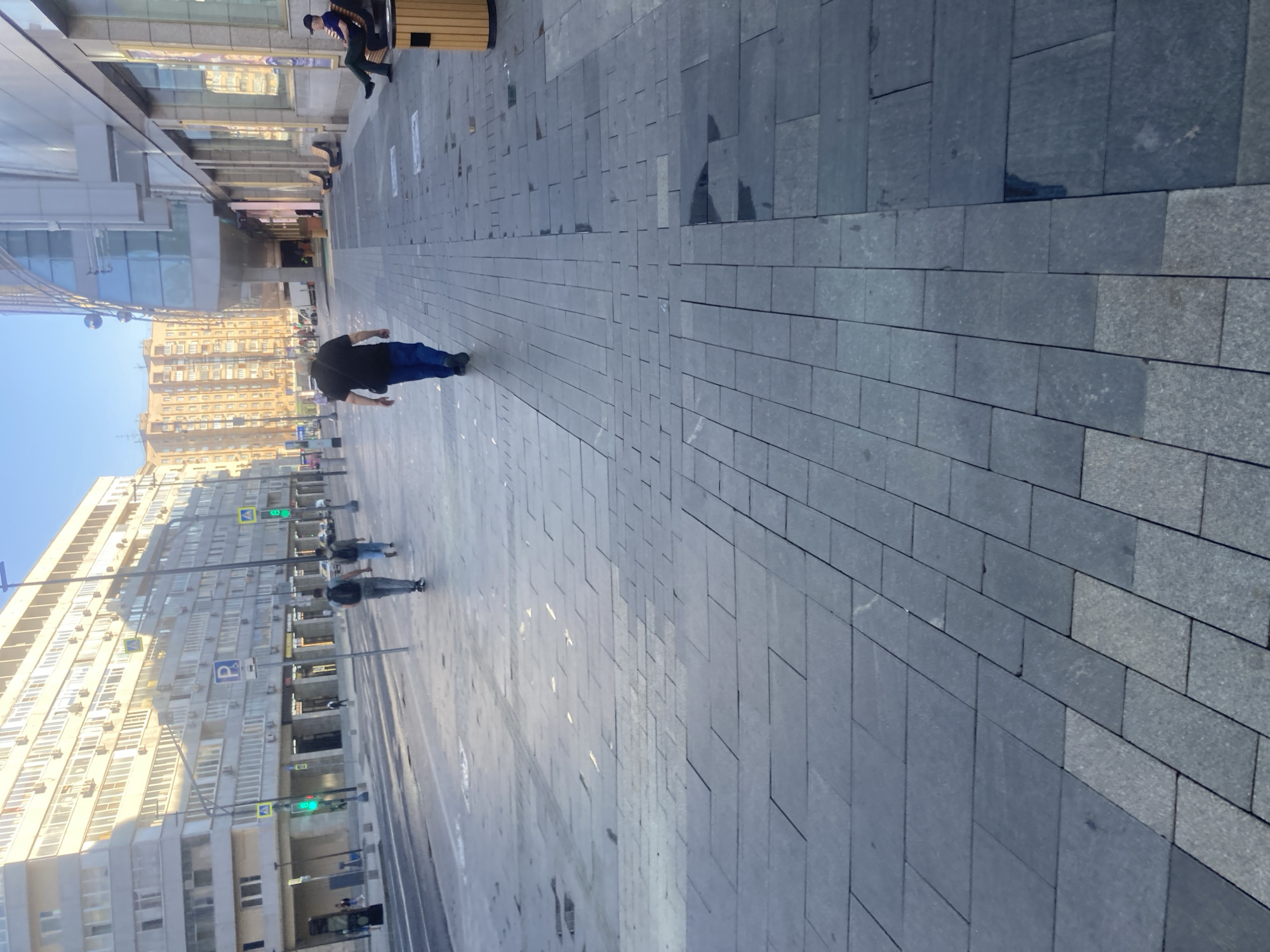 The image shows a street scene in Moscow, Russia. The photograph is taken at a low angle, causing distortion. A wide, paved walkway made of rectangular stones dominates the foreground. The walkway is relatively clean and stretches from the right of the image towards the left. A person in dark clothing is walking along the walkway. They appear to be wearing a dark top and blue pants. There are other people, with some blurred by motion, walking along the street. On the left side of the image are several tall buildings with numerous windows. Traffic lights and street signs are visible in the background. The sky is clear and blue. The overall impression is of a modern, urban environment.