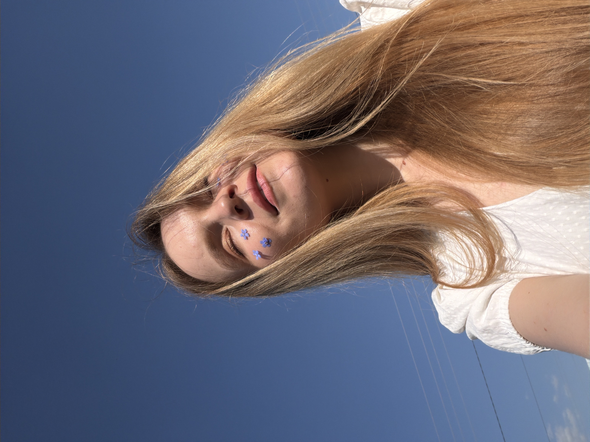 A woman with long blonde hair is lying on her back in Kadryakovo, Russia, looking directly up at the camera. She is wearing a white top. Three small blue flowers are placed on her left cheek. The background is a clear, bright blue sky. Power lines are faintly visible in the lower right corner.