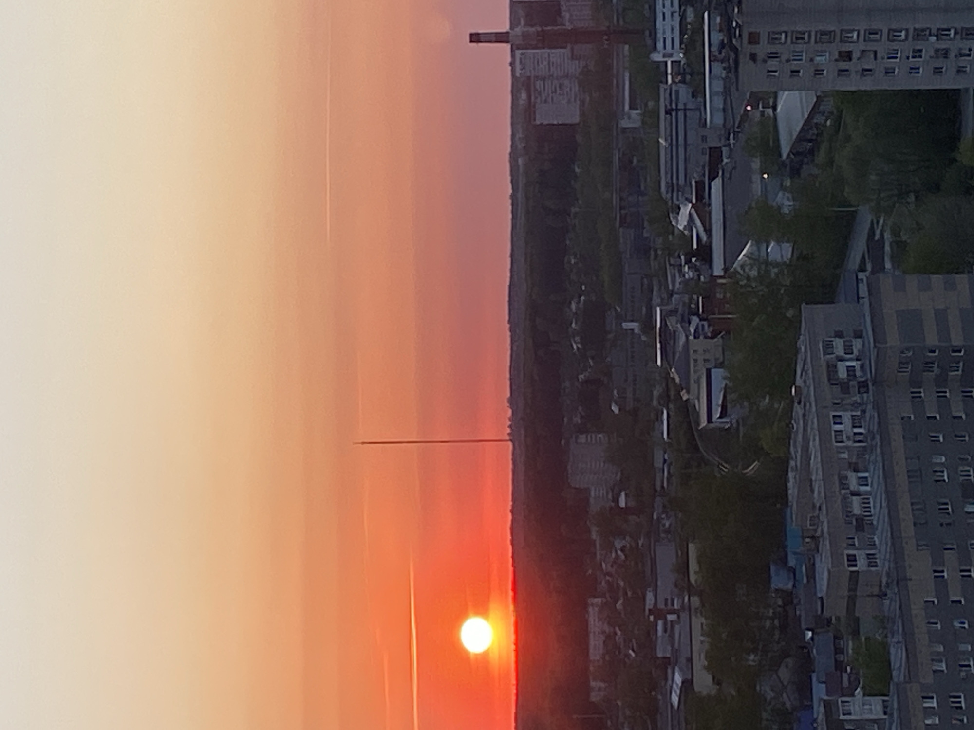 The image shows a sunset over the city of Izhevsk, Russia. The sun is low on the horizon, creating a vibrant orange and red glow. A tall structure, possibly a television tower, is silhouetted against the sunset. The city is spread out below, with buildings of varying sizes and styles visible. Trees are interspersed throughout the urban landscape. The sky is clear.