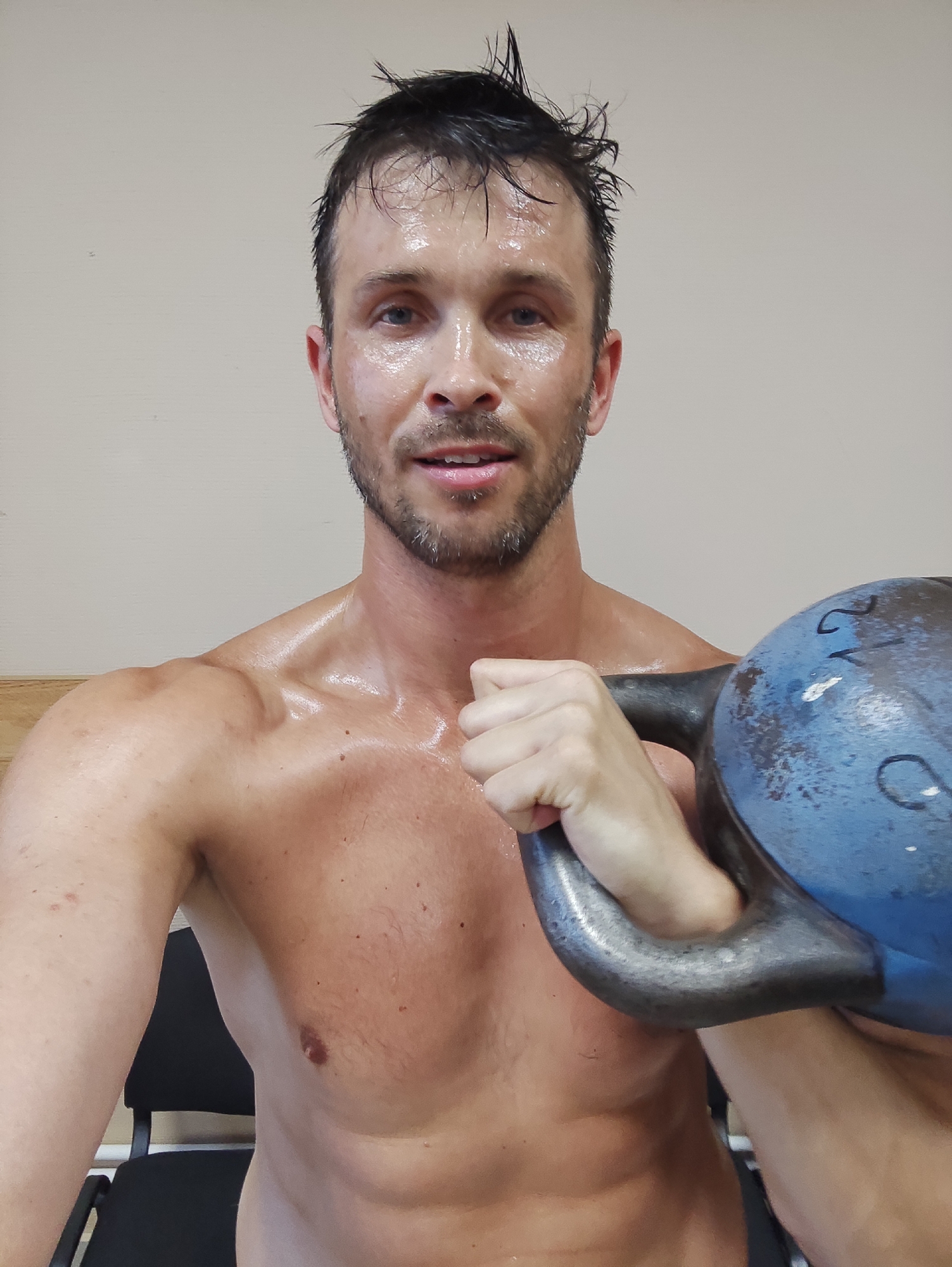 A man is pictured from the chest up, holding a blue kettlebell with a black handle. He has sweat on his face, neck, and shoulders, and his hair is wet and styled upwards. He is looking directly at the camera with a slight smile. The background is a plain, light-colored wall. The man is wearing a wooden plank that is visible in the lower left corner of the image. The scene appears to be indoors, possibly in a gym or workout space.