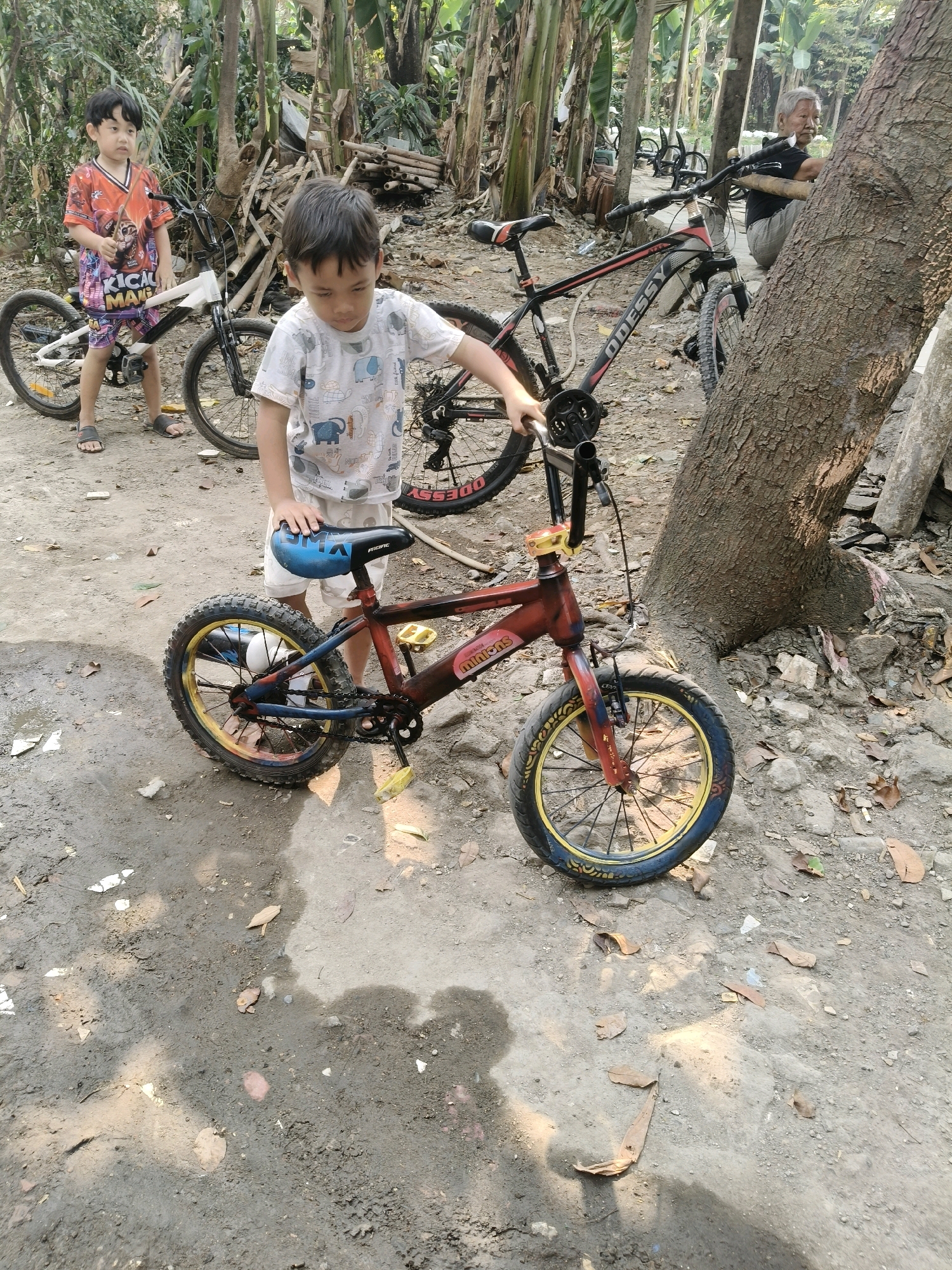 Two young boys are in an outdoor, dirt-covered area with trees and other bicycles. The boy in the foreground, wearing a light-colored t-shirt with an elephant pattern and white shorts, is standing next to a small red and yellow bicycle. He is holding the bicycle seat with his right hand. The bicycle has black tires with yellow rims and a blue seat. The boy in the background, wearing an orange and red patterned t-shirt and patterned shorts, is standing to the left of a white bicycle. He is holding a stick or pole in his right hand. In the background, an elderly person is seated next to a large tree. The environment appears to be a residential area or yard in South Tangerang, Indonesia, indicated by the presence of banana trees and the tropical vegetation. The ground is a mixture of dirt, sand, and scattered leaves.