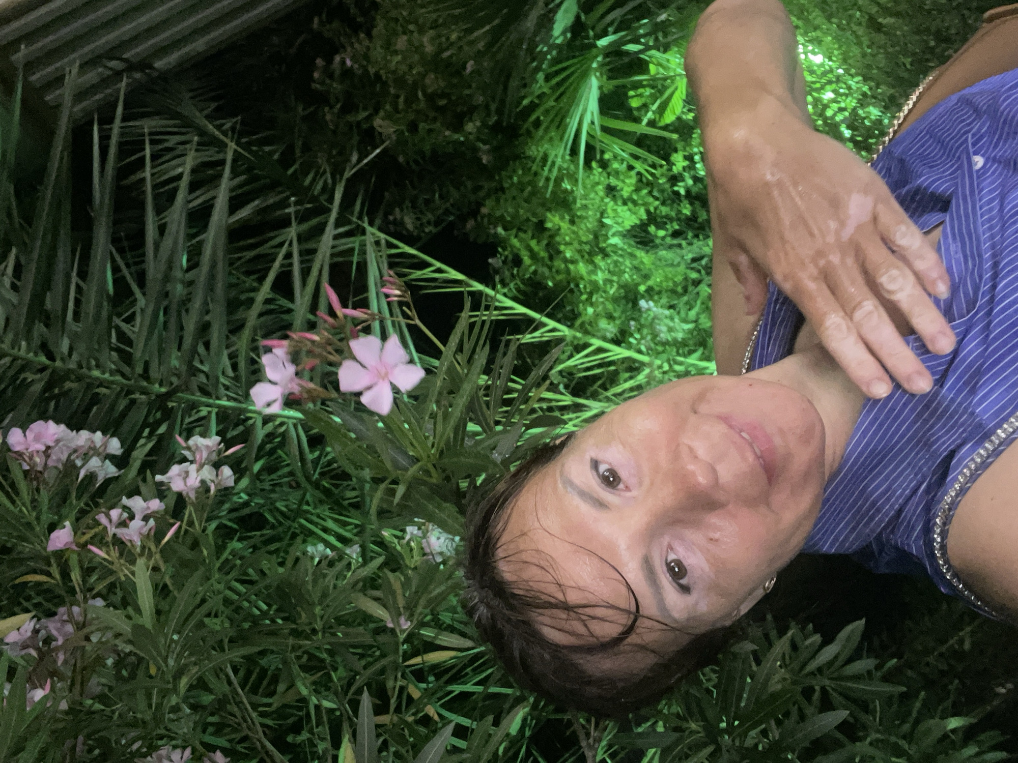 A woman with short, dark, wet hair and light skin is taking a selfie outdoors at night, surrounded by lush greenery. She is wearing a blue and white striped shirt with a silver decorative trim. Her right arm is raised, with her hand resting on her chest. Her skin appears to have a patchy, lighter tone in some areas. The background is dominated by dark green foliage, illuminated by bright green artificial lighting. Some light pink flowers are visible among the leaves. The overall impression is that of an outdoor setting, possibly a garden or park, within the city of Sochi, Russia.
