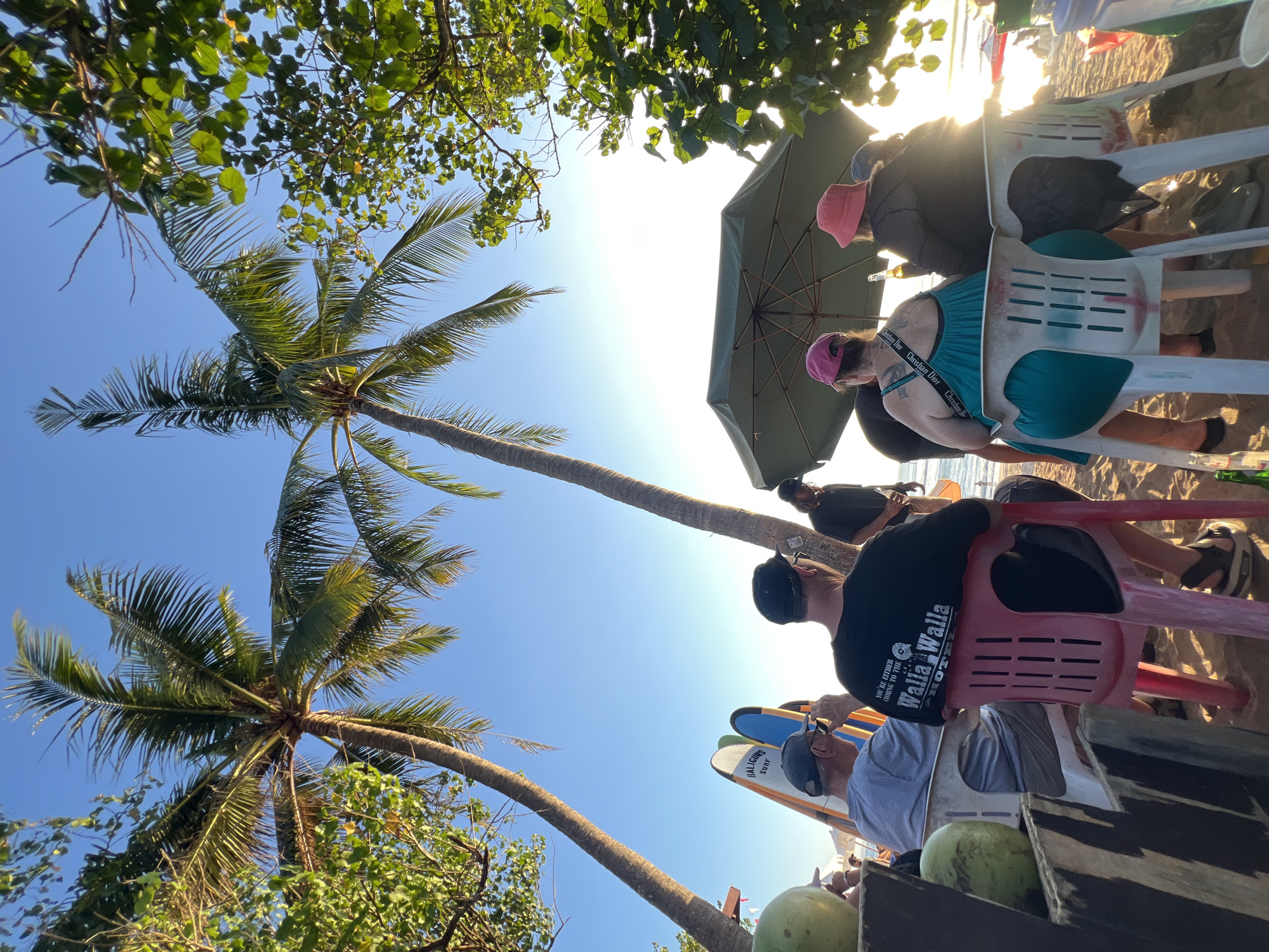 In Kuta, Indonesia, on a sandy beach with palm trees and lush foliage, a group of people are seated on chairs. Two tall palm trees frame the scene, with their fronds reaching towards a clear blue sky. In the foreground, several people are seated, facing away from the camera, under a large green umbrella. One individual wears a black t-shirt with "Walla Walla Hotel" printed on it. Another person wears a teal dress and a pink hat. Two surfboards stand upright in the background, with text visible on one that reads "BALIGUNS SURF". Two green coconuts are visible on the left side of the frame. The sun is low in the sky, creating a warm glow and casting long shadows on the sand. The overall atmosphere suggests a relaxed setting, possibly a beachside cafe or bar.
