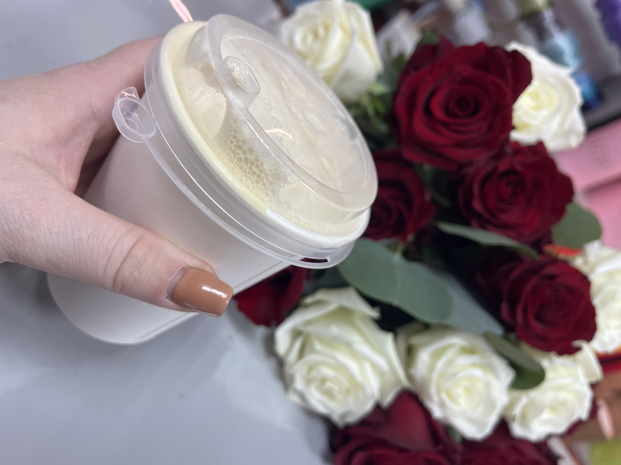A hand is holding a white plastic cup with a clear lid, likely containing a beverage, against a backdrop of red and white roses. The scene is set on a grey surface, possibly a table. The person's fingernail is painted a tan color. The location is Chaikovsky, Russia.