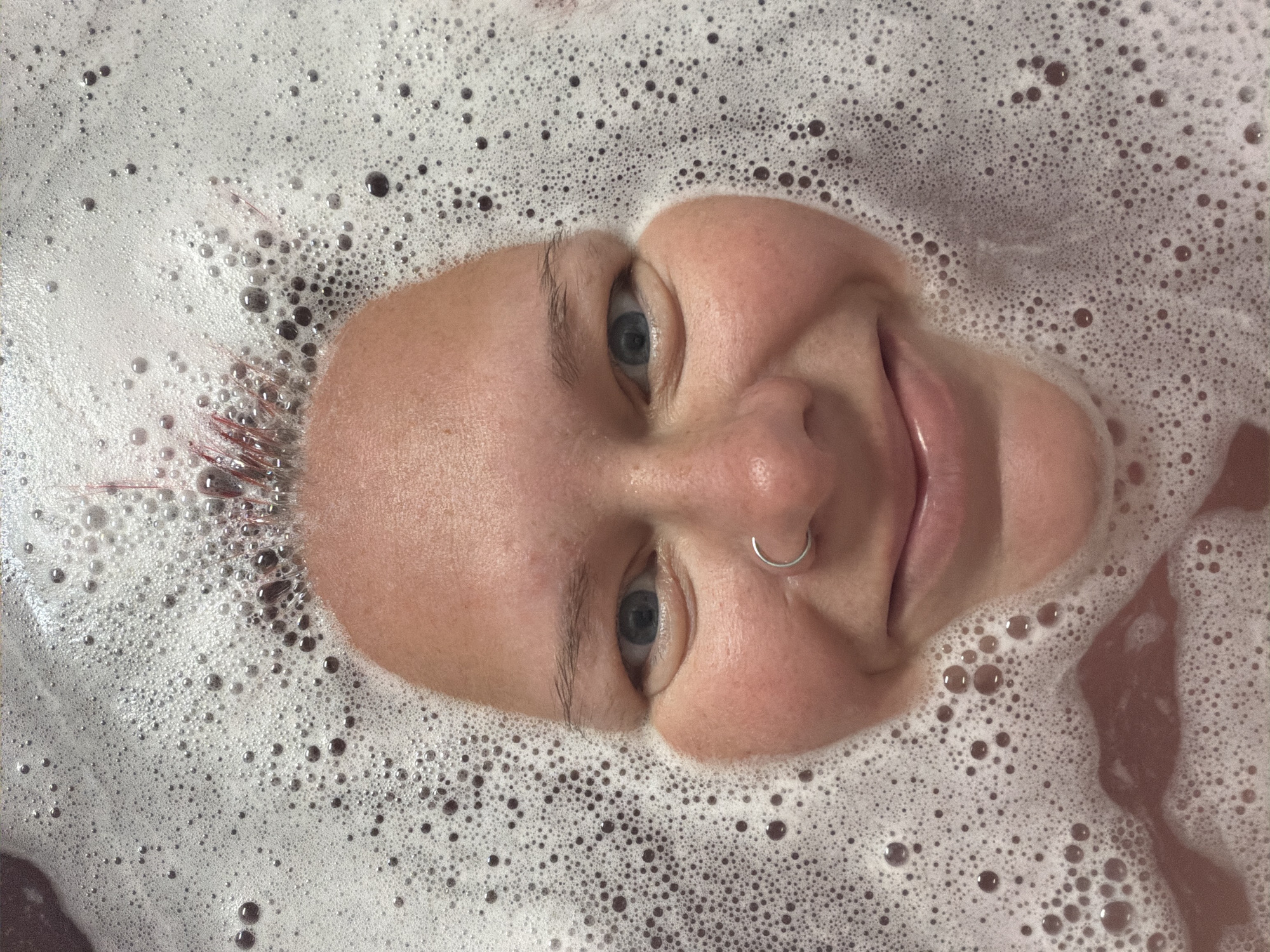 The image shows a close-up, high-angle view of a person's face submerged in a bathtub filled with pinkish water and covered in white foam. The person's eyes are open and looking at the camera, and they have a nose ring. Their mouth is also visible, with lips slightly parted in a smile. The foam surrounds their face, creating a bubble-like frame. The background is out of focus, but appears to be a bathroom setting. There is no information about the location in the city of Chaikovsky, Russia.