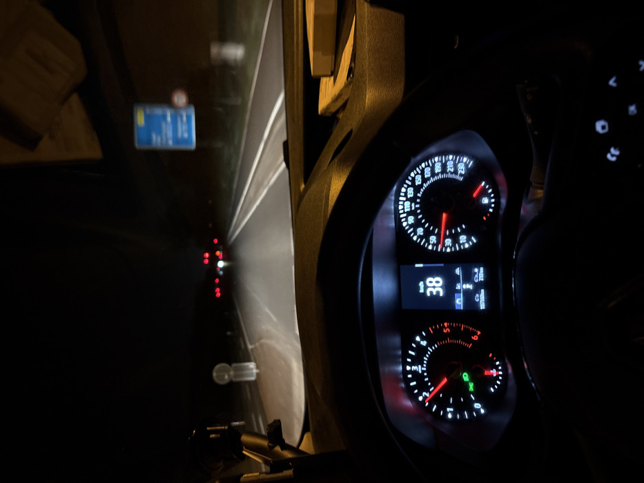 The image presents a forensic view from inside a vehicle at night, driving on a road in the vicinity of Ratingen, Germany. The lower half of the frame is dominated by the vehicle's dashboard and instrument cluster. The digital speedometer centrally displays "38 km/h," consistent with the analog speedometer's needle position. The tachometer shows approximately 1500-2000 RPM. Several indicator lights are illuminated, including the headlights and a fuel warning light. A partial view of the steering wheel is visible at the bottom. An object, possibly a device mount, is affixed to the dashboard on the left, and a light-colored item rests on the dashboard to the right.
Through the windshield, the vehicle's headlights illuminate a two-lane asphalt road with clear white lane markings. On the left side of the road, a white circular sign with a red border is partially visible. On the right, a blurry blue rectangular road sign with white text is discernible, indicating directions to "Ratingen," "Düsseldorf," and "Duisburg," with "Ratingen" listed as 7 km away. Below this, a circular sign with a red border, likely a speed limit sign, is partially visible. Distant red tail lights of other vehicles are visible further down the road, indicating traffic. The environment is dark, suggesting an unlit road section, possibly bordered by roadside vegetation.