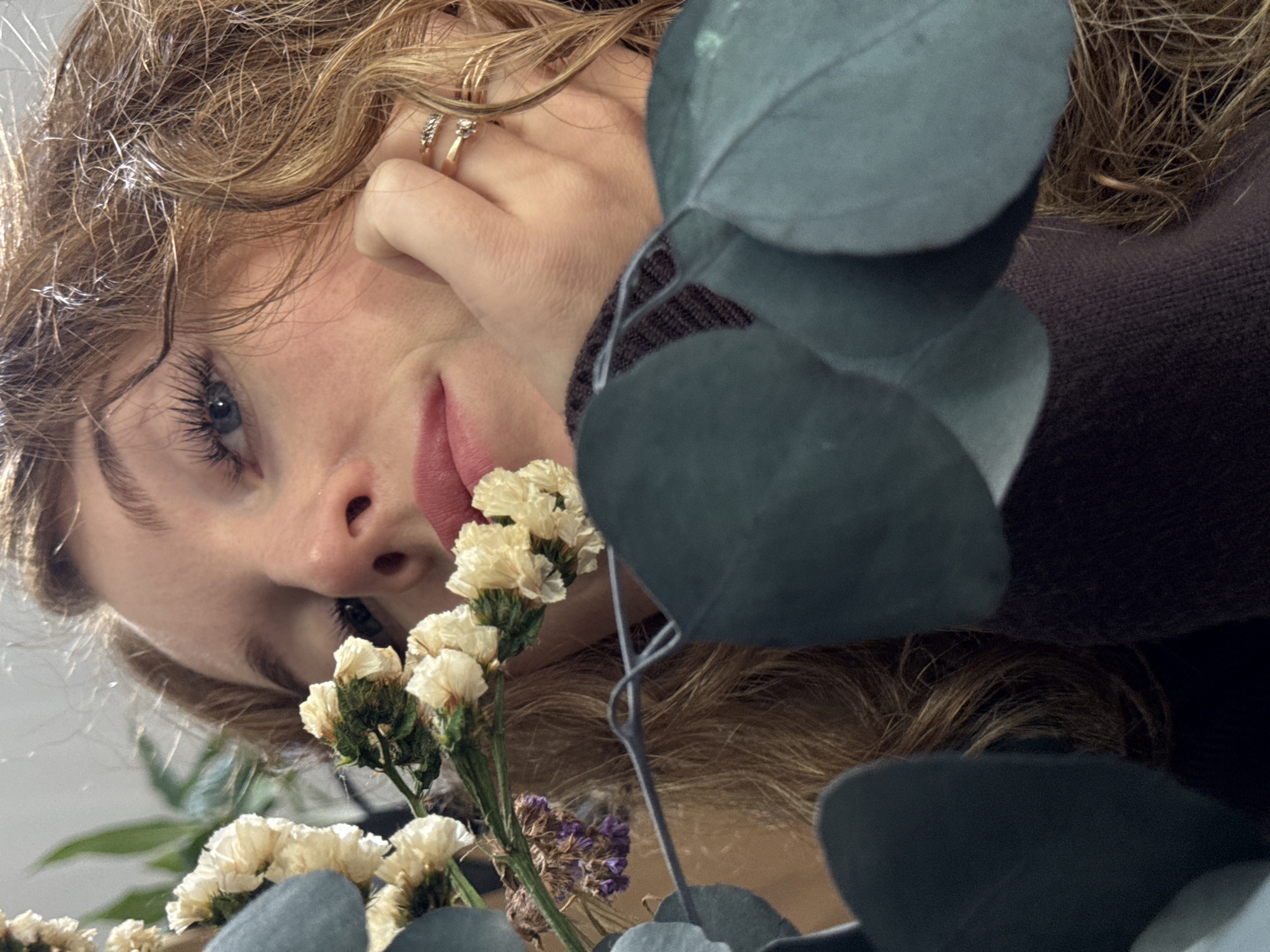 The image shows a person with fair skin, light brown curly hair, and blue eyes. Their face is tilted towards the camera. They are wearing rings on their fingers and a dark brown sweater. A bouquet of dried white and purple flowers and eucalyptus leaves is positioned in front of the person, partially obscuring the view. The photograph has a close-up perspective, focusing on the person's face and the floral arrangement. The location is not discernible from the image.