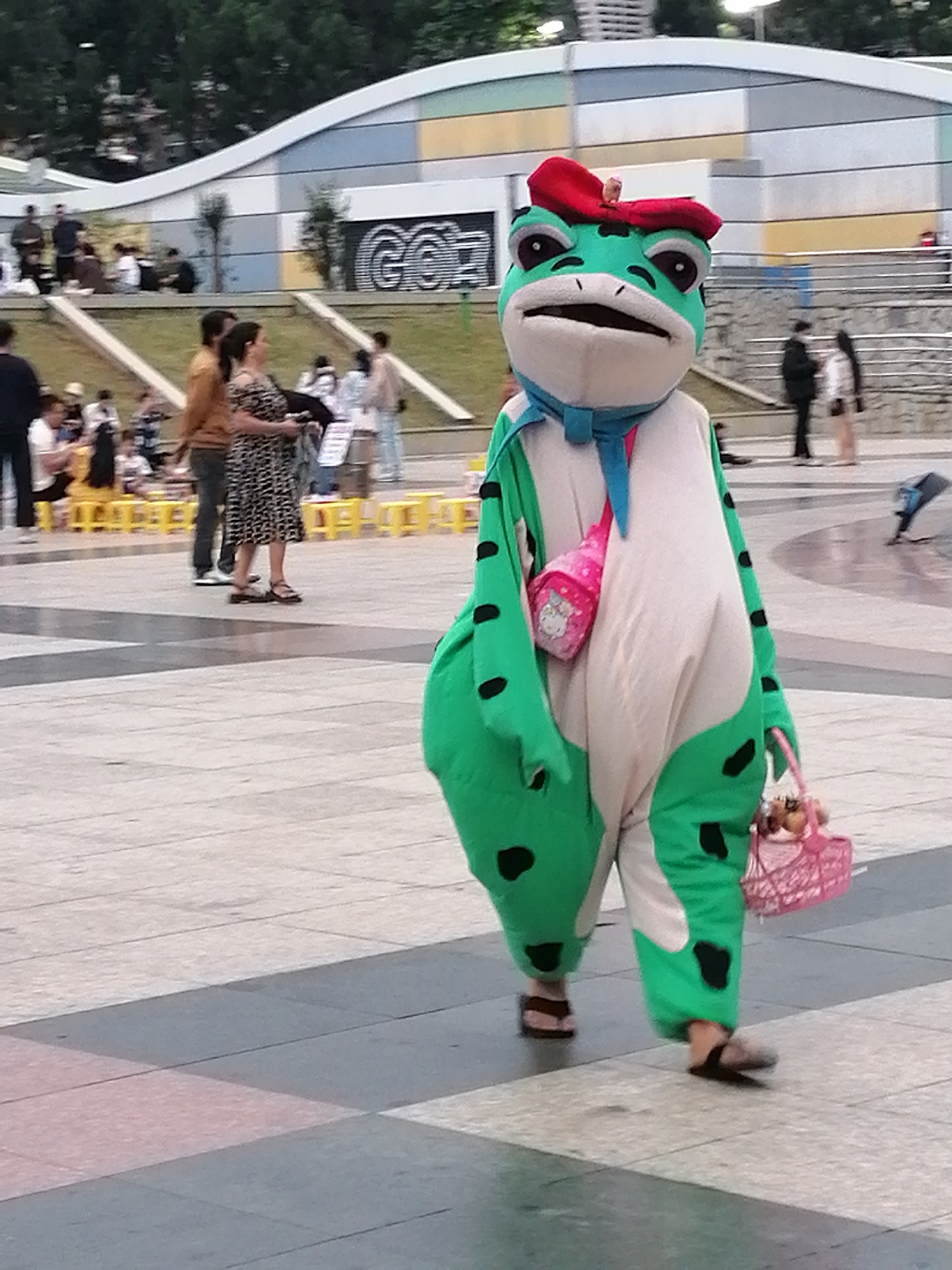 In Da Lat, Vietnam, a person in a large, green frog costume with black spots walks on a paved area. The costume features a red bow on its head, a blue scarf, and a pink Hello Kitty purse slung across its chest. The frog is also carrying a pink basket with items inside. In the background, there are several people, some sitting on yellow chairs, and a building with a curved, colorful facade. The environment appears to be an outdoor public space, possibly a park or plaza, with trees visible in the distance.