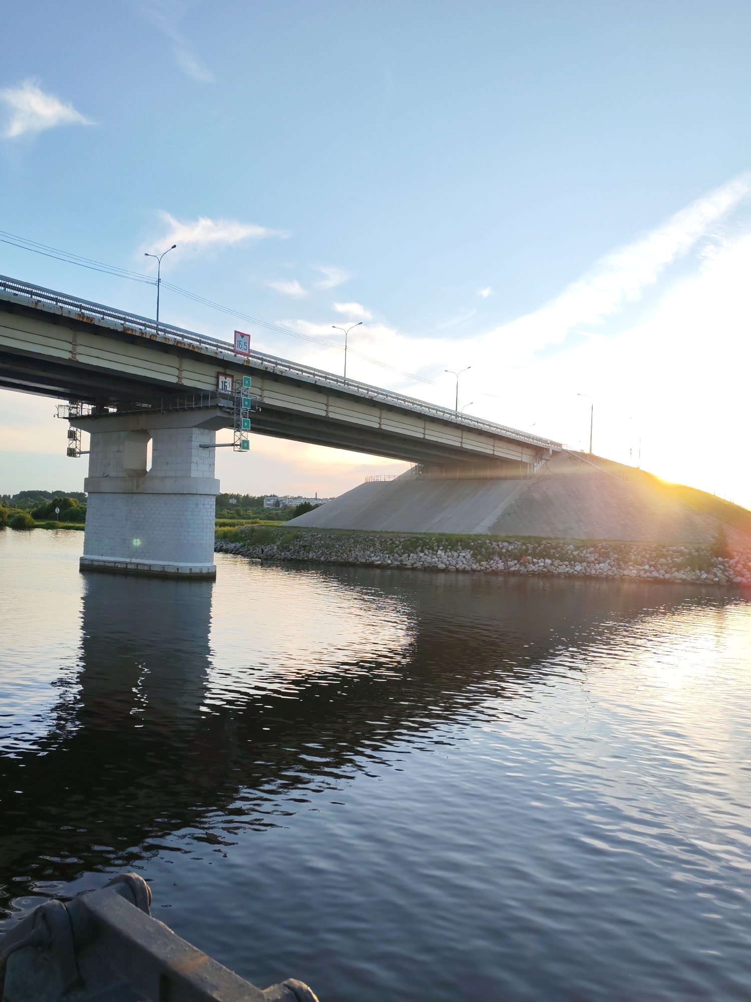 This is a photograph of a large concrete bridge spanning a body of water. The bridge has a wide, flat deck supported by substantial concrete pillars. The sun is setting or rising, casting a warm golden light and long shadows across the scene. The sky is a clear blue with some wispy clouds. The water is calm, reflecting the bridge and the sky. The embankment leading up to the bridge is covered with rocks and grass, with some trees and buildings visible in the distance on the left side. The overall mood is peaceful and serene, typical of a sunset or sunrise over a natural landscape. There is some visible text on the bridge: a sign that reads "16.5" within a red circle, and a smaller sign below it with green and white symbols. These likely indicate height or weight restrictions for vehicles. The location is Sheksna, Russia.