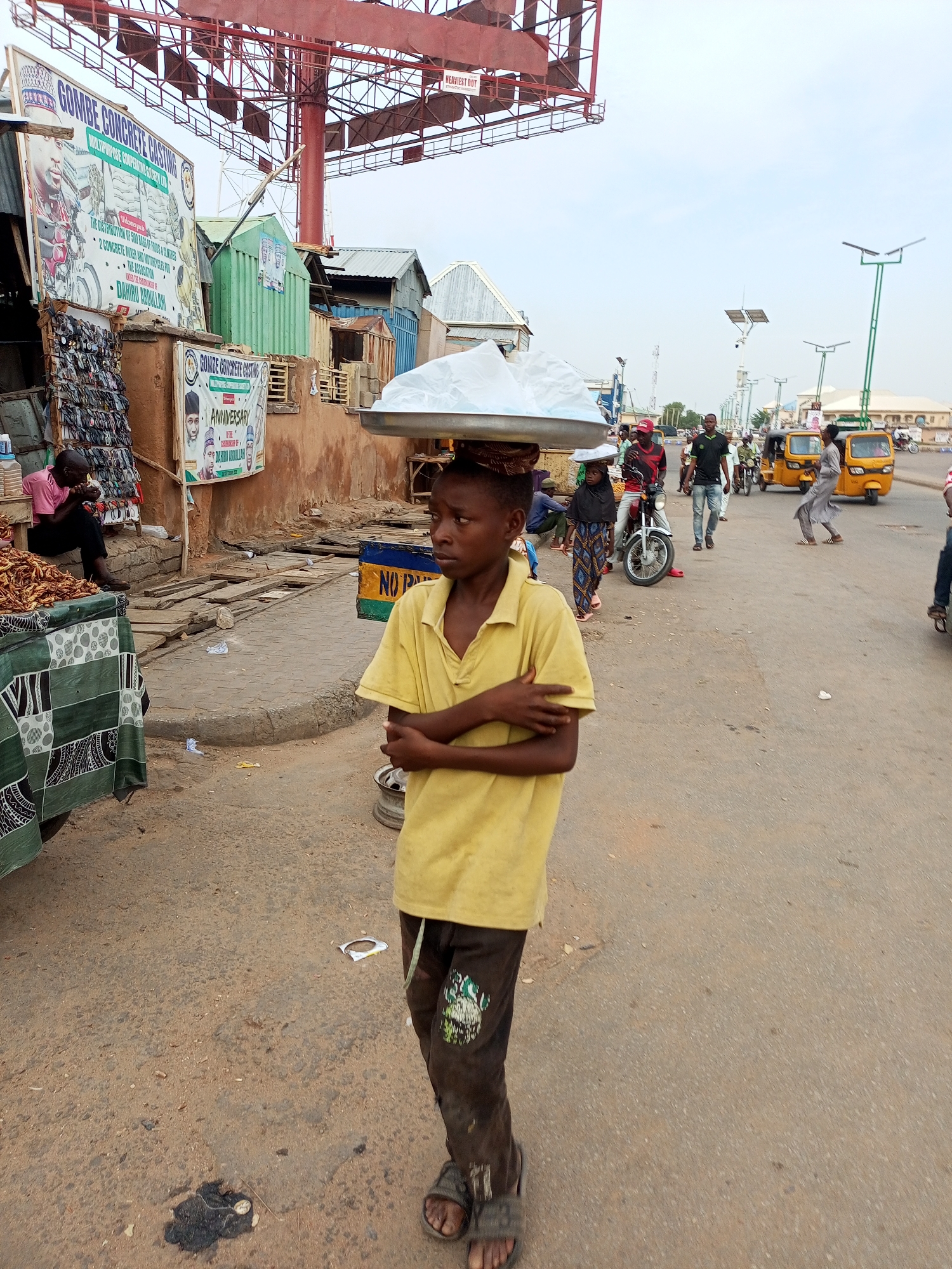 A young boy wearing a yellow polo shirt and dark pants walks with his arms crossed, carrying a large silver platter on his head. The platter is covered with a white sheet, obscuring its contents. The boy is standing on a dirt and asphalt road in what appears to be a marketplace in Arawa, Nigeria. In the background, there are various market stalls, some made of corrugated metal and wood, and a large billboard with text in English and what appears to be local script. Several people are visible in the background, some walking, some on motorcycles, and three yellow auto-rickshaws are parked on the side of the road. The environment suggests a busy, outdoor commercial area with elements of both traditional and modern infrastructure.