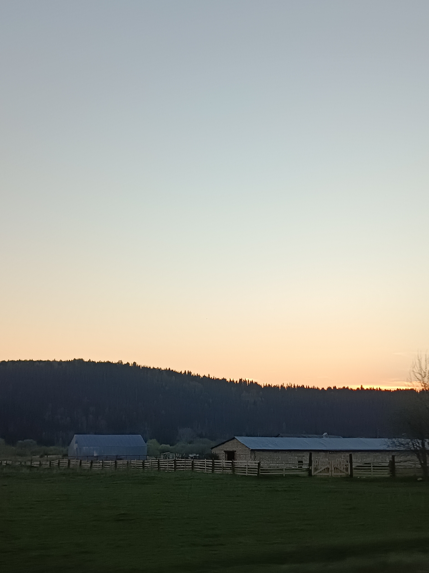 The image shows a rural landscape in Ust-Kishert, Russia, at sunset. A dark, densely forested hill forms the horizon. In the foreground, a grassy field stretches towards a wooden fence. Two barns are visible behind the fence: one with a gray roof and walls and the other barn is stone or masonry construction with a silver roof. The sky transitions from pale blue to orange near the horizon.