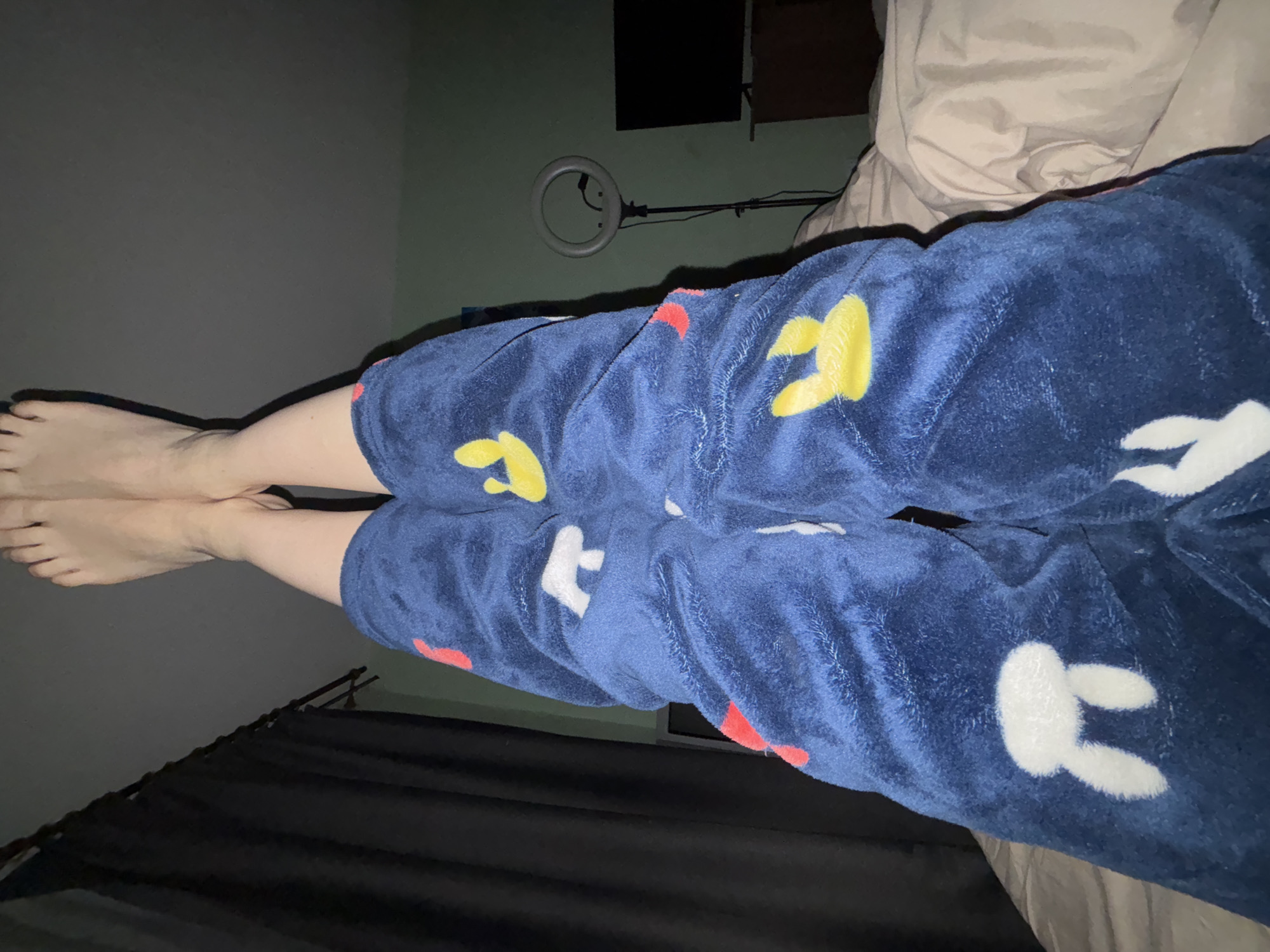The image shows a person lying on a bed, wearing blue plush pajama pants with cartoon rabbit designs in white and yellow. The person's bare feet are visible at the edge of the frame. A black curtain is visible in the bottom left corner. A ring light is visible on a stand in the background. The wall is a pale green color. The image was taken in an indoor environment, possibly a bedroom. The location of Murino, Russia cannot be determined from the information provided.