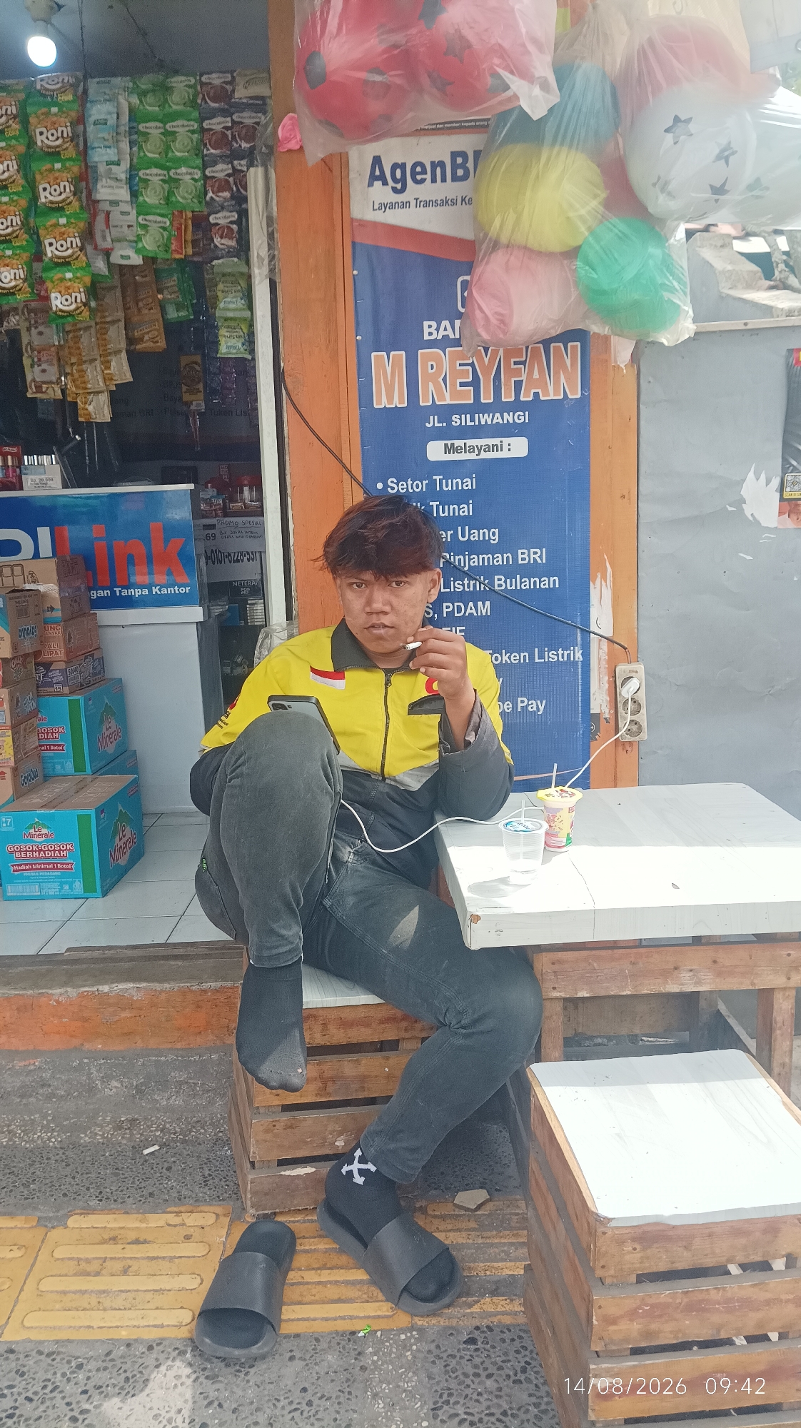 The image depicts an outdoor scene in Tasikmalaya, Indonesia, outside a small shop. A young man, appearing to be in his late teens or early twenties, is seated on a wooden stool. He is wearing a yellow and black jacket, dark gray jeans, black socks, and black slides. He is holding a lit cigarette in his right hand and a smartphone in his left. A white table is in front of him, with two drinks on it, one in a clear plastic cup and another in a yellow and red cup. Behind the man, the shop has shelves stocked with various food and drink items, as well as a blue banner advertising services from "AgenBR" and "M REYFAN". The shop appears to be located on a street with a yellow tactile paving strip and a grey concrete surface. The overall impression is of a casual, everyday moment captured outside a local business.