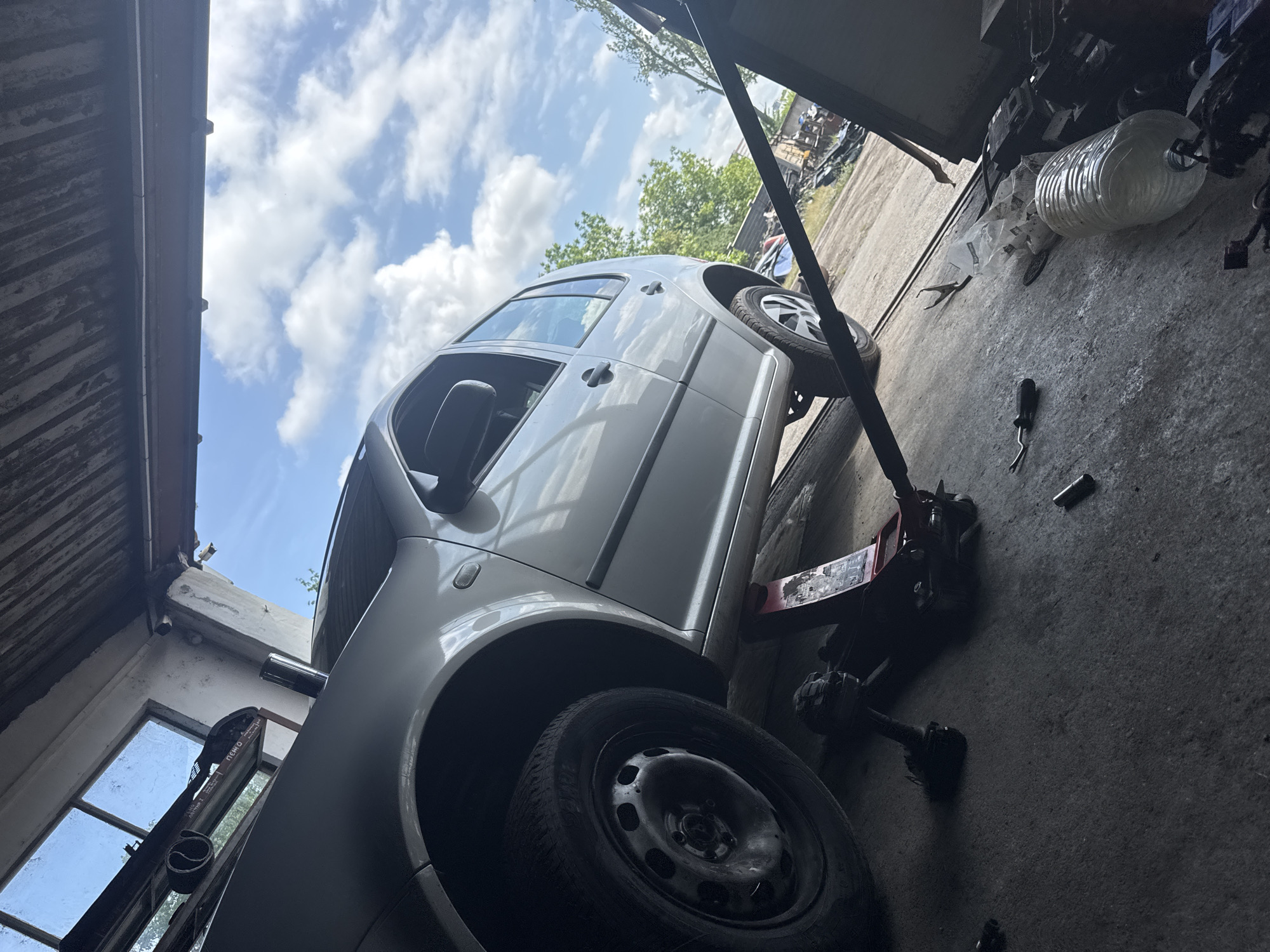 The scene depicts a silver car, a Volkswagen Golf, elevated on a red hydraulic jack inside what appears to be an auto repair shop or garage. The front left wheel has been removed. The garage has a concrete floor littered with tools, a plastic water bottle, and other debris. Natural light is coming in through a window on the left and a doorway on the right, revealing a cloudy sky outside. The location is implied to be Yambol, Bulgaria. There are no people visible in the image.