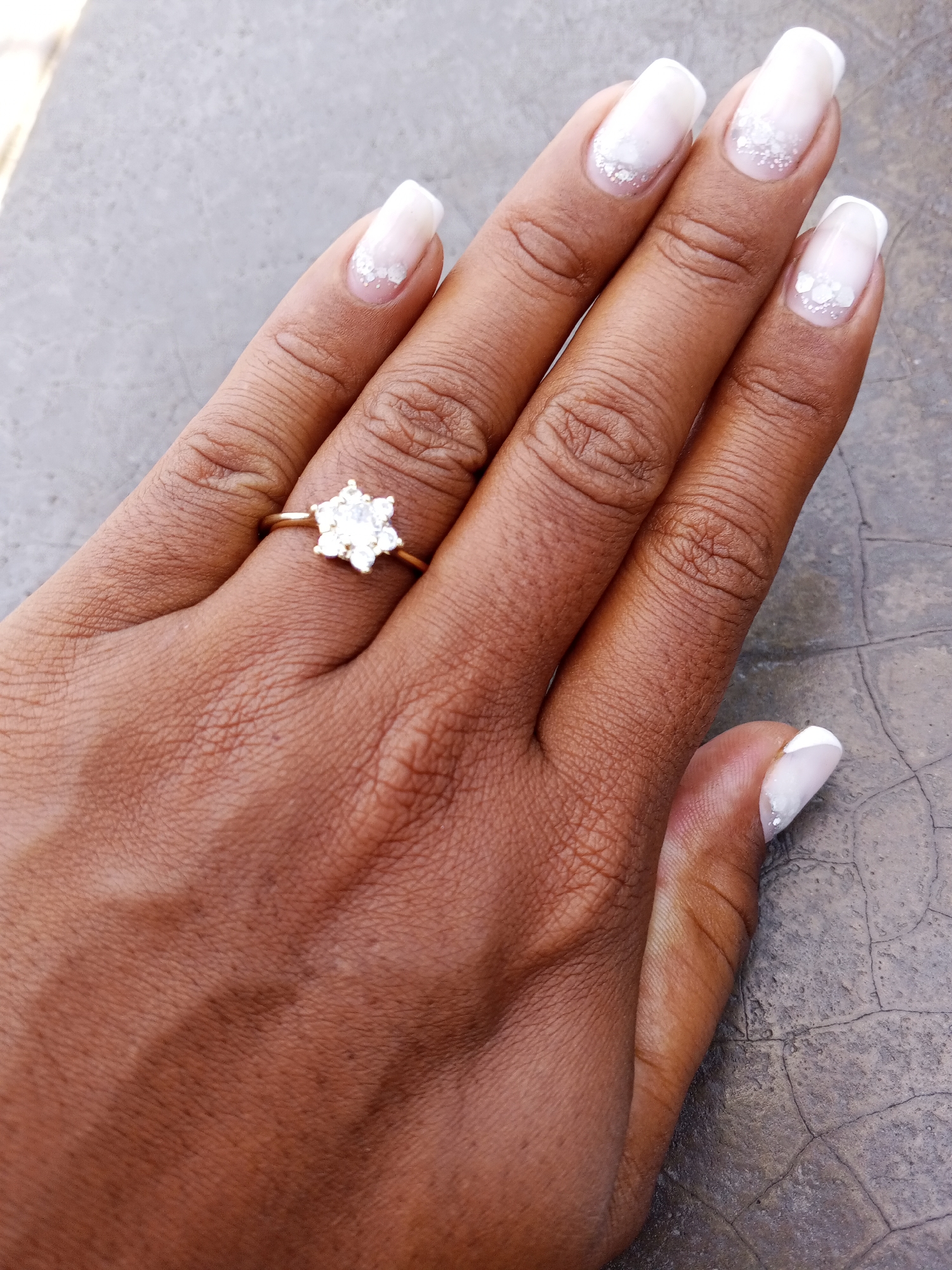 A close-up shot depicts a hand adorned with a gold ring featuring a white, flower-like stone. The ring rests on the ring finger. Each fingernail is painted white with a translucent base, adorned with white flower-like details near the base. The hand is situated on a grey, cracked surface. The city and location are not discernible from the image.