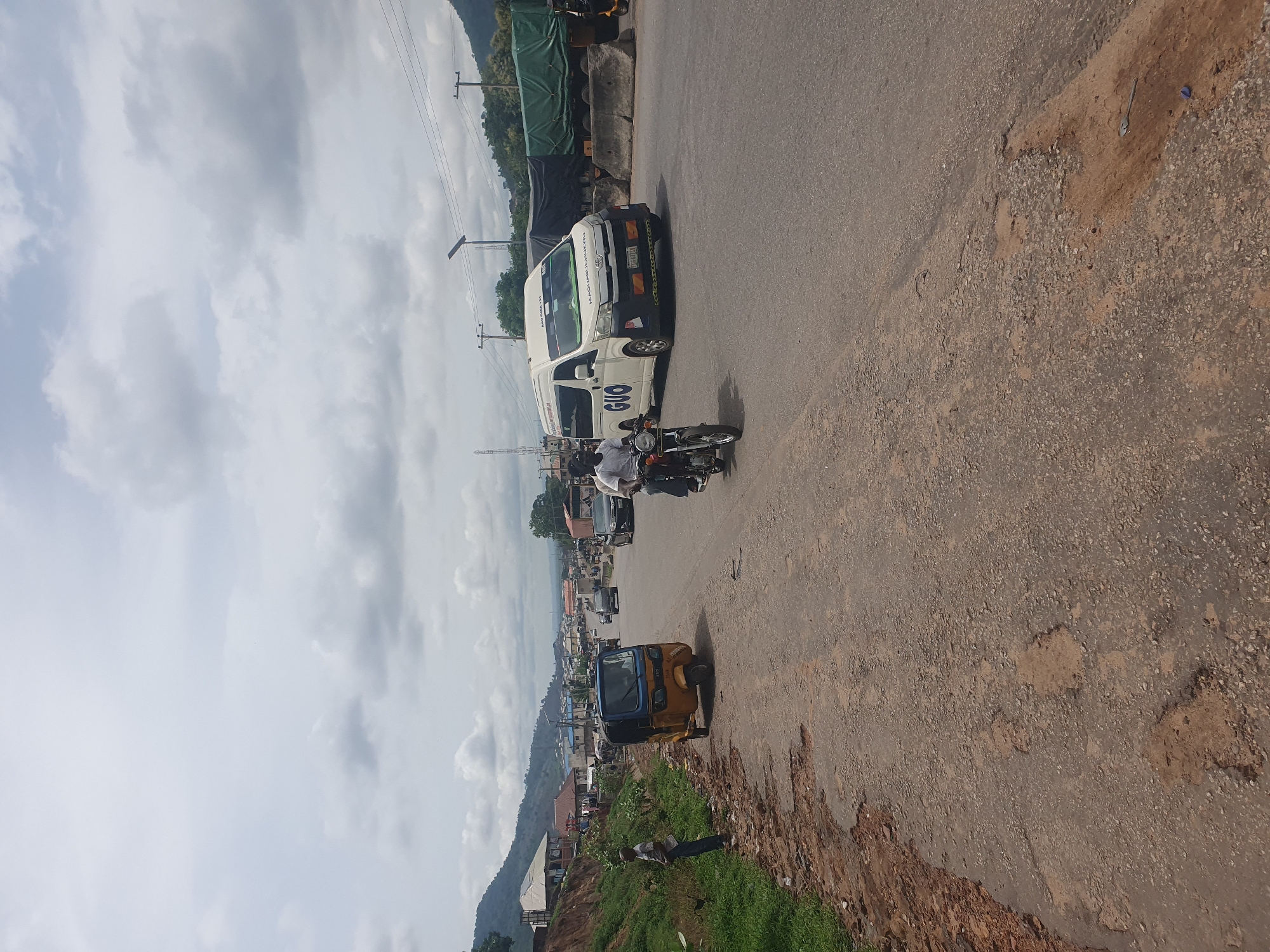 The image shows a street scene in Lokoja, Nigeria. A white van with "GUO" markings is driving on the road, followed by a motorcycle carrying two people. In the foreground, a yellow tricycle is parked on the side of the road, near an area of eroded earth. A person is standing nearby on the grassy embankment. Other vehicles and buildings can be seen further down the road. The sky is cloudy.