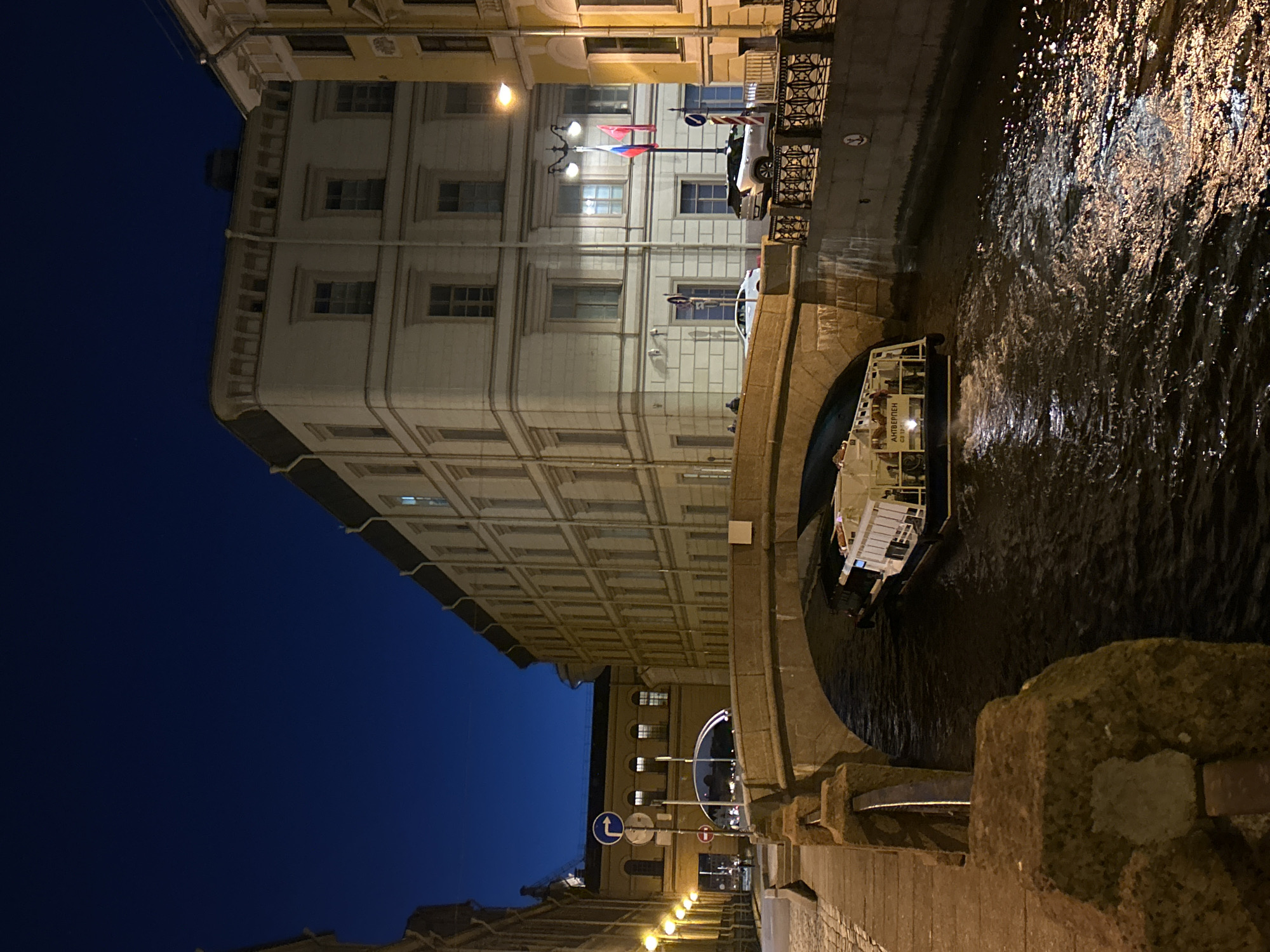 An eye-level photograph shows a nighttime cityscape of Saint Petersburg, Russia, featuring a canal, a bridge, and illuminated buildings. The camera is positioned looking down at the water to the right and the tall buildings on the left. The dark blue sky is in the upper left corner. A boat is passing under the bridge. The water in the canal is dark with light reflecting off the surface. A stone bridge spans the canal in the middle of the scene. The stone bridge is on the right and is made of stone blocks. A stone block is in the lower right foreground.
The architecture on the left is ornate, with multiple rows of windows. Russian flags can be seen on one of the buildings. The lights from the building are illuminated. A series of streetlights lead into the distance.