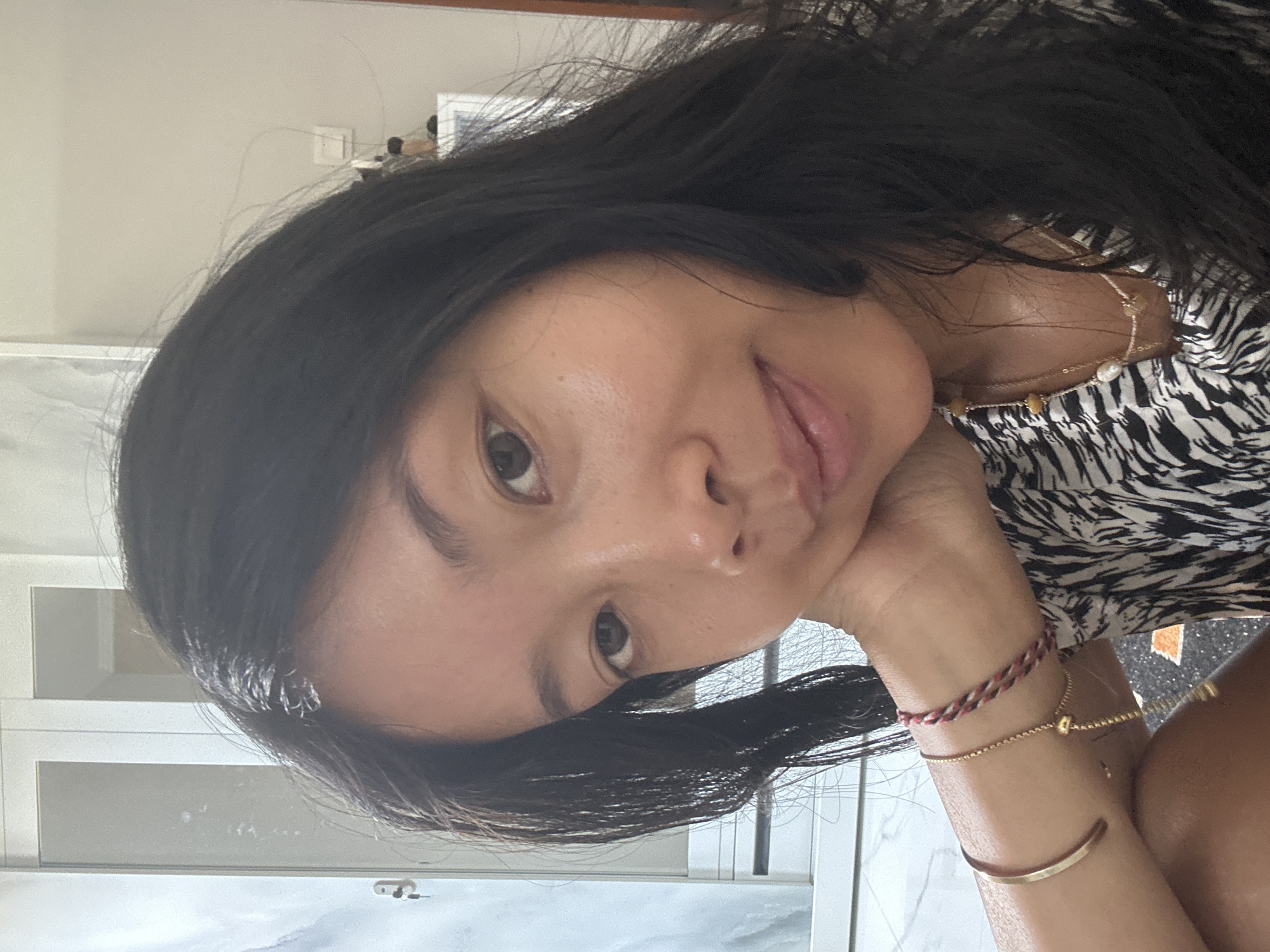 A young woman of Asian descent with dark hair is captured in a close-up, waist-up selfie. She is resting her chin on her left hand, which is adorned with several bracelets, including a gold cuff and a braided red, white, and black bracelet. Her gaze is directed towards the camera with a slight smile. She is wearing a black and white patterned top. The background suggests an indoor setting with a closet or wardrobe visible to the left and a shelf with some items to the right. The image appears to be taken in Sukawati, Indonesia.