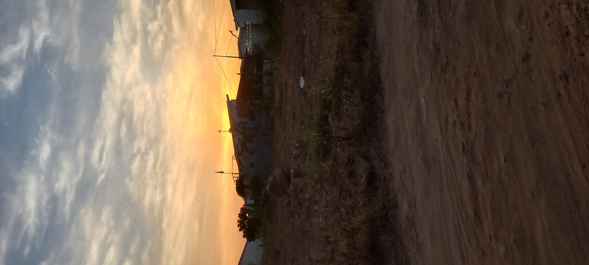 The image depicts a rural landscape in Loa Lepu, Indonesia, during a sunset. The sky is filled with a gradient of colors, transitioning from a bright orange near the horizon to a pale blue at the top, with scattered clouds. In the mid-ground, several buildings with unfinished roofs are visible, along with some vegetation. A large rock sits in the foreground, surrounded by dry grass and dirt. The ground in the foreground is a dirt path with tire tracks. There are no people visible in the image.