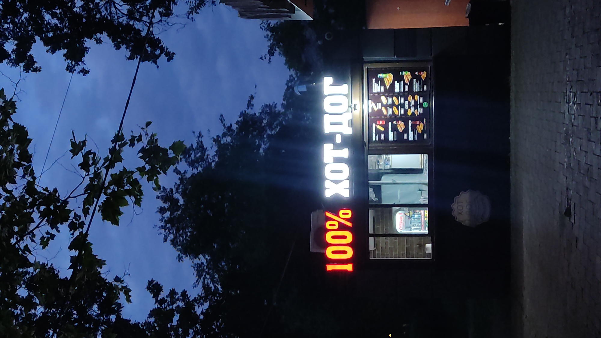 A neon sign displaying "100% ХОТ-ДОГ" illuminates a small food kiosk at dusk. The sign is divided into two parts: "100%" in red neon and "ХОТ-ДОГ" in white neon. The kiosk has a window where customers can order, and a menu board is visible inside, showcasing various hot dog options. Outside the kiosk, there is a paved area. The background consists of dark trees and a cloudy sky. The overall atmosphere is that of a typical street-side eatery operating in the evening. The food kiosk is located on Dzerzhinskogo Street, Krasnodar, Russia, a commercial area with a mix of residential and retail establishments.