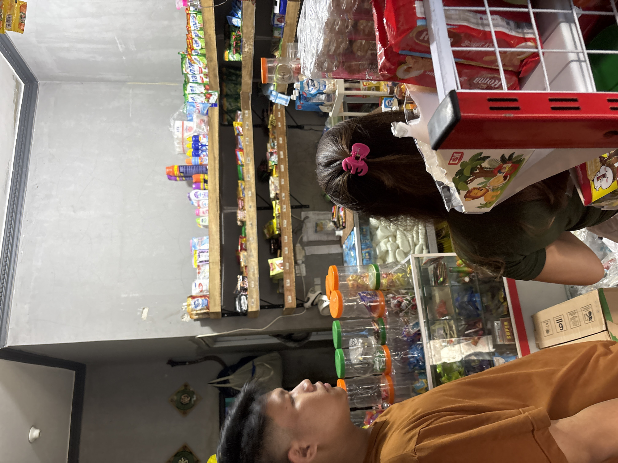 In a dimly lit convenience store in North Jakarta, Indonesia, a young man with dark, spiked hair stands in profile, wearing an orange t-shirt. He is looking towards the right side of the frame. To his right, a woman with long, dark hair tied with a pink clip, faces away from the camera, her torso visible as she also looks towards the right. They appear to be in an aisle of the store.
The store's interior features shelves stocked with various products. On the left, clear plastic containers filled with colorful candies are stacked on top of a display cabinet. The cabinet itself contains more small packaged items. Above these, wooden shelves are organized with rows of packaged goods, including small bottles, boxes, and bags. One shelf has a variety of colorful bottles and cans, some with blue and orange labels. Another shelf displays rows of brightly colored packets. Towards the back of the store, more shelves are visible, some with larger white bags of products.
The walls are painted a neutral gray, with some visible texture and discoloration. A white ceiling light is visible in the upper left corner. In the foreground, a red display rack holds red packaged items, and a cardboard box with graphics of animals and trees is placed on a lower shelf. The overall impression is of a small, local shop filled with everyday consumer goods.