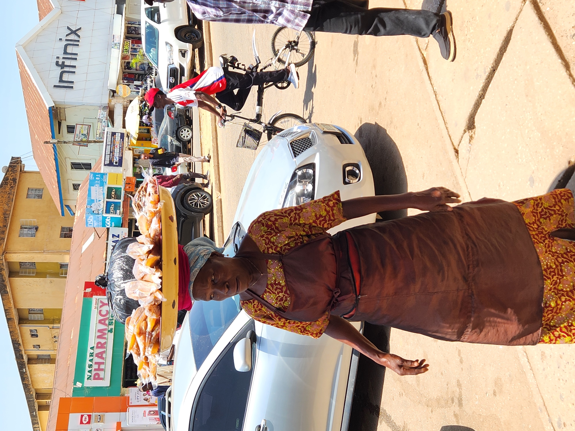 The image depicts an elderly woman selling bread on a street in an urban area. She is wearing a patterned top, a brown apron, and a headscarf, and is standing next to a white car. She is carrying a tray of bread. Behind her, there is a pharmacy called "NASARA PHARMACY & Ste" and various advertisements including "Itel," "MTN," and "CITY 200". A building with the sign "Infinix" is visible in the background, as well as other vehicles, including a pickup truck and a person on a bicycle. There are pedestrians and a bicycle in the background. The location appears to be a busy street with commercial establishments.