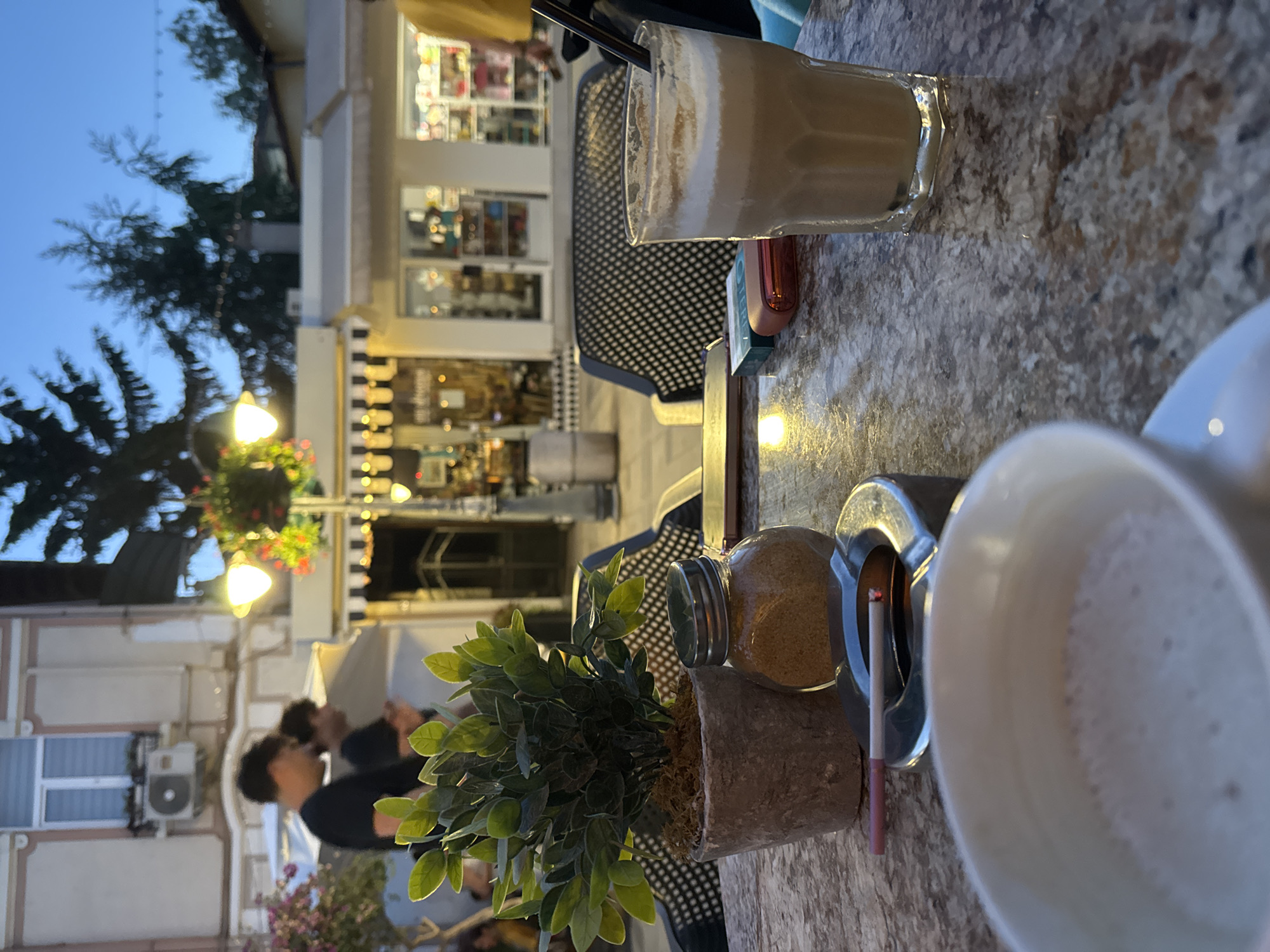 The image depicts an outdoor seating area at a cafe on Bogoridi Street in Burgas, Bulgaria, during twilight. In the foreground, a marble-patterned table holds a white cup of coffee with foam, an ashtray with a lit cigarette, a small jar of brown sugar, and a potted artificial plant. To the right, a tall glass contains a frothy, light-brown beverage with a black straw. Behind the table, black outdoor chairs are visible. In the background, the facade of buildings with shops and awnings line the street, illuminated by streetlights and lights from within the establishments. Two young men are walking in the distance, one in a black t-shirt and the other in a darker shirt. The overall atmosphere suggests a leisurely evening at a cafe in a city setting.