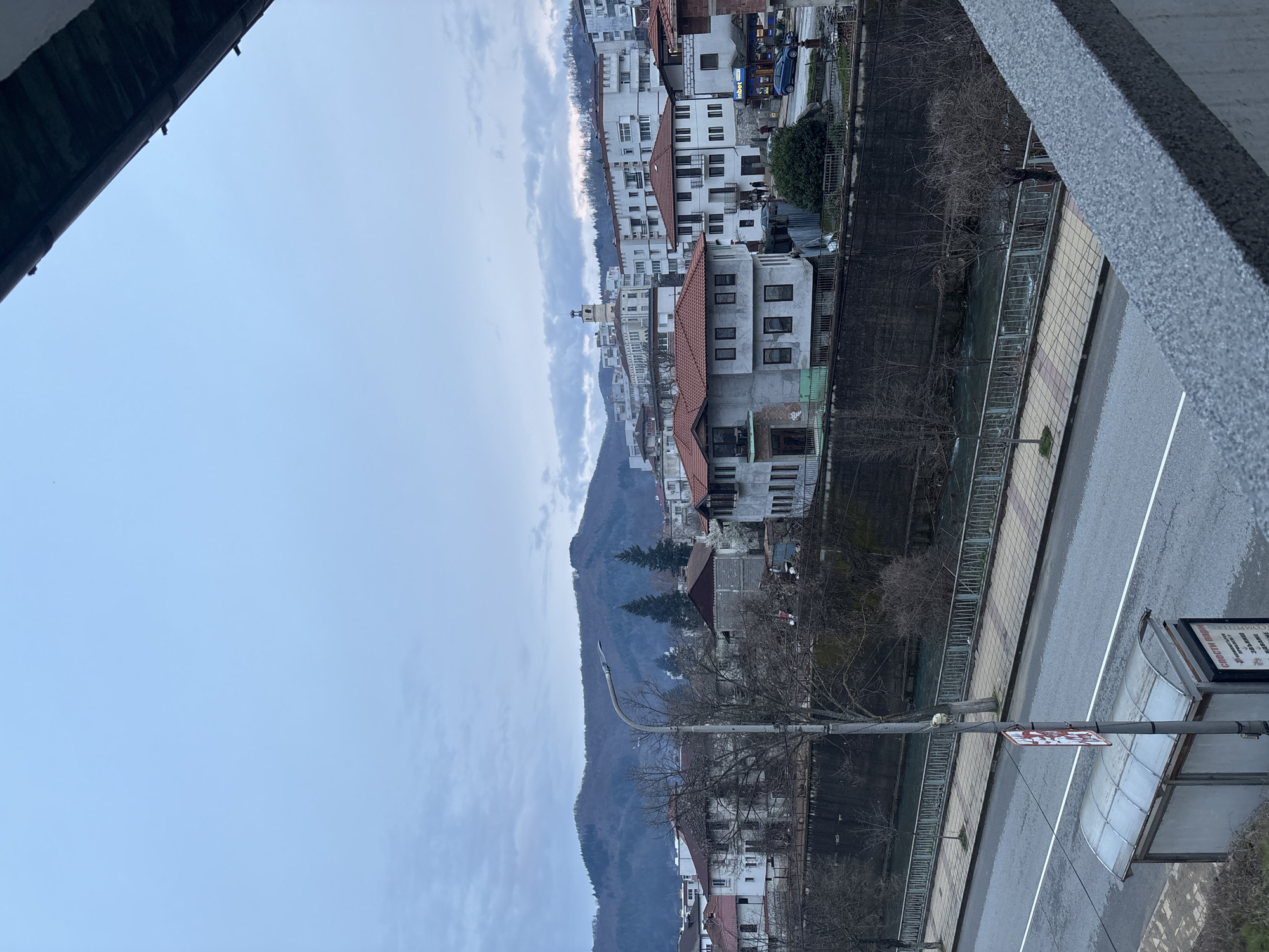 View from an elevated position in Zlatograd, Bulgaria. A grey mountain rises behind a row of white buildings with red roofs. A river flows between the buildings and a street, which includes a bus stop sign and shelter. The sky is overcast. Balcony structure partially visible in the foreground. No people are visible.