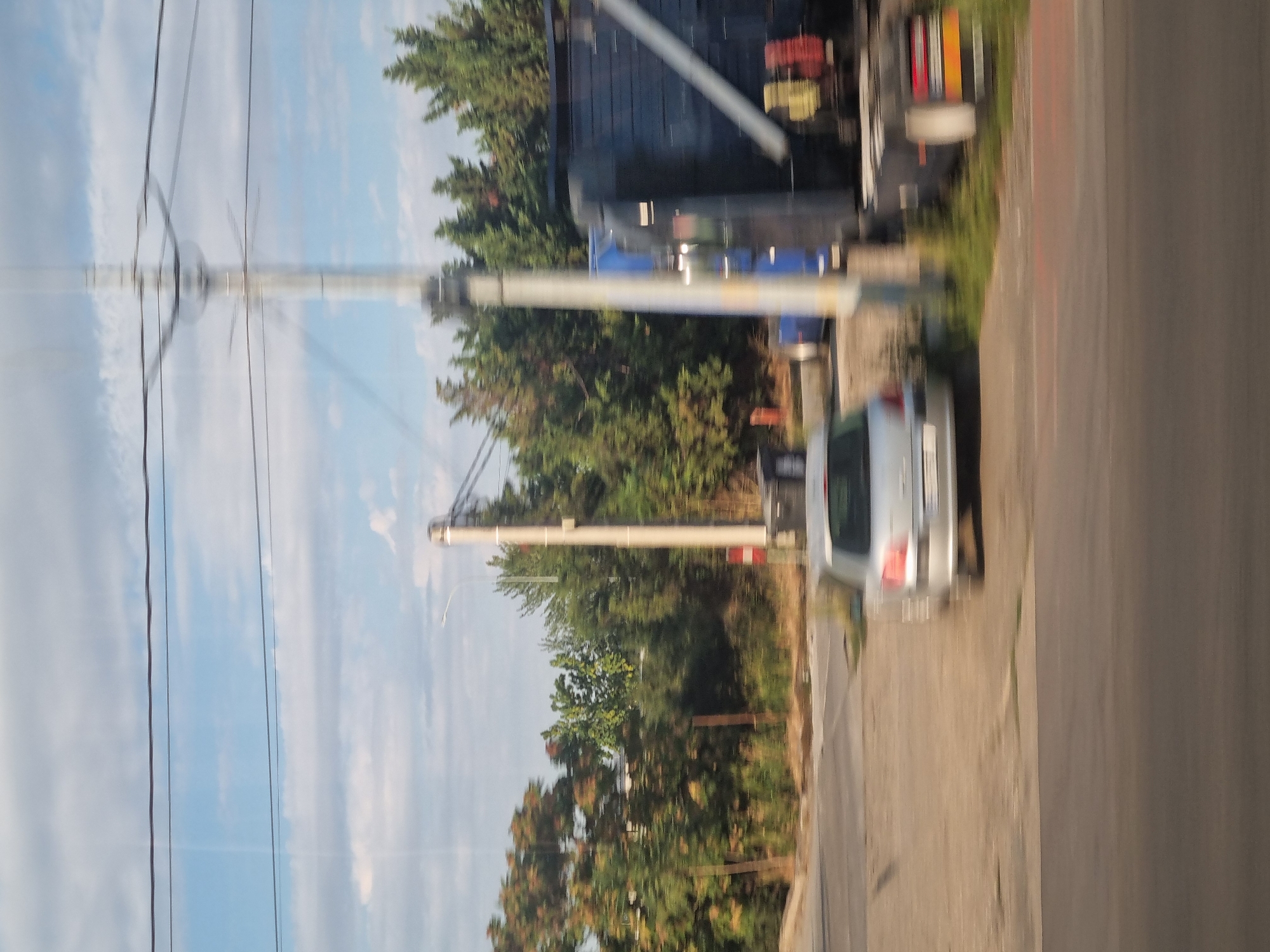 This is a daytime, outdoor scene, likely taken from a moving vehicle, depicting a street environment in Sântion, Romania. In the foreground, a gray car is parked or moving slowly on a paved surface. Behind it, a large, dark-colored truck with some red and yellow markings is visible, along with a blue trailer. A tall, utility pole with multiple wires extends vertically through the image, and several trees with green foliage line the background. The sky is partly cloudy with a light blue hue. The overall impression is that of a street or roadside view in a semi-urban or rural setting.