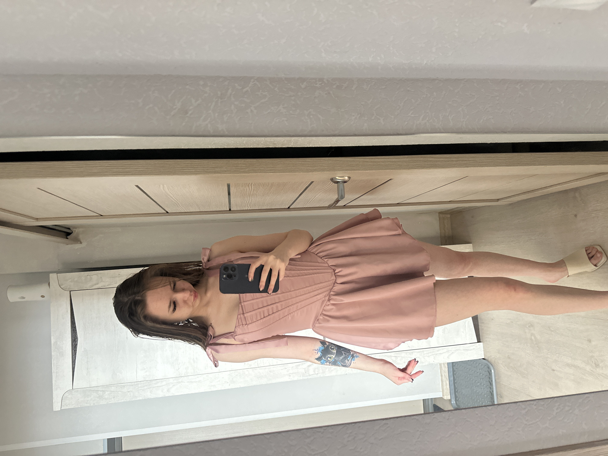 A person is reflected in a mirror. The person is holding a smartphone. The person is wearing a pink dress and beige shoes. The environment is indoors. The location is in the city of Unknown, Unknown.