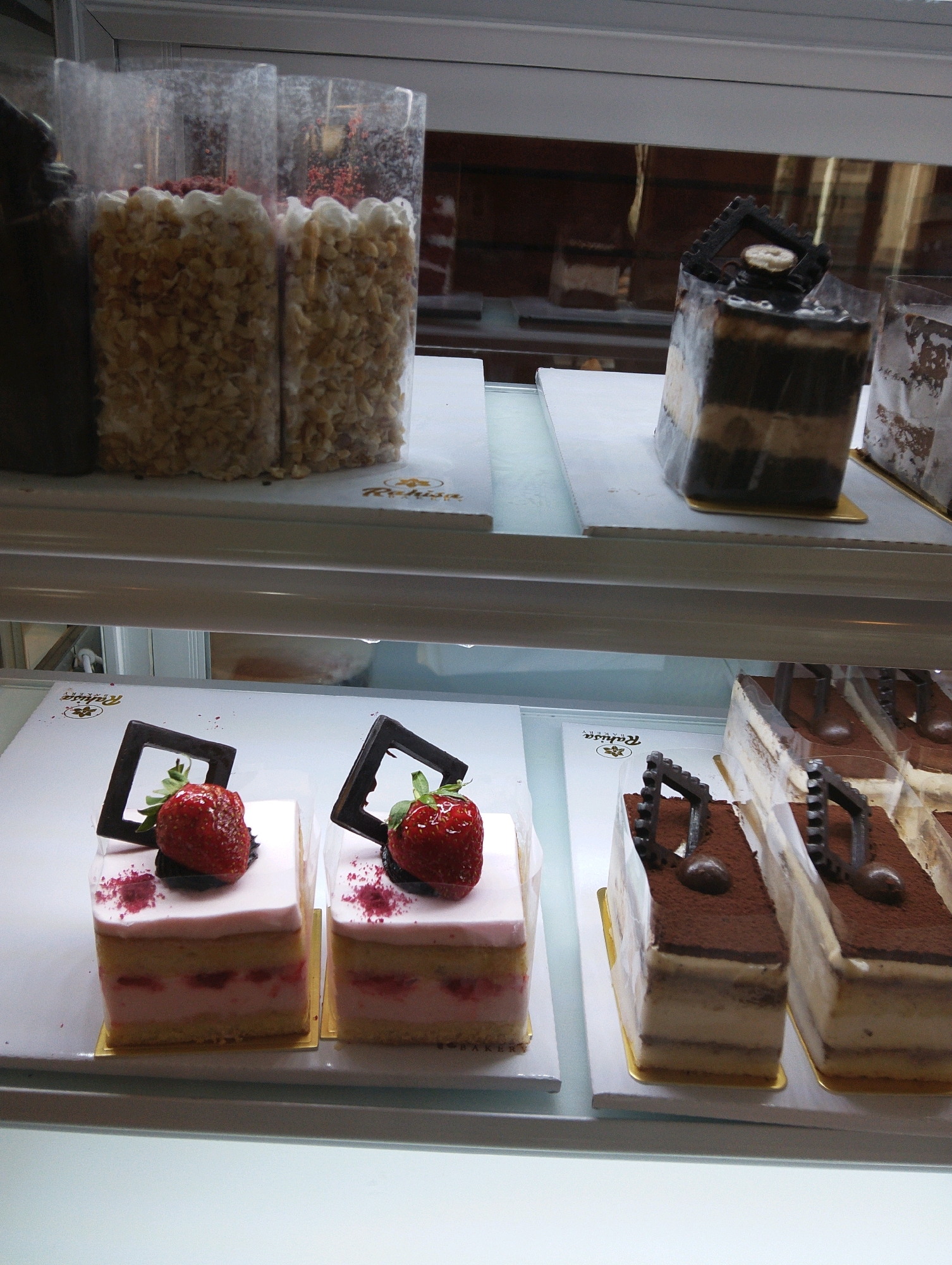 The image displays a refrigerated display case at the "Rakusa" bakery in Manaruwi, Indonesia, filled with various pastries. In the top shelf, two large plastic containers are filled with a granular, light-colored confection, likely candied nuts or cereal pieces, with hints of red topping. To the right, a rectangular chocolate cake with layered dark and light brown frosting is visible. The middle shelf showcases two individual square cakes topped with a strawberry and dark chocolate garnish, with a light pink frosting layer. Next to these, there are several slices of tiramisu cake, each dusted with cocoa powder and topped with a dark chocolate decoration. The bottom shelf is mostly empty, with a white surface reflecting the light. The overall environment is a clean and well-lit bakery display.