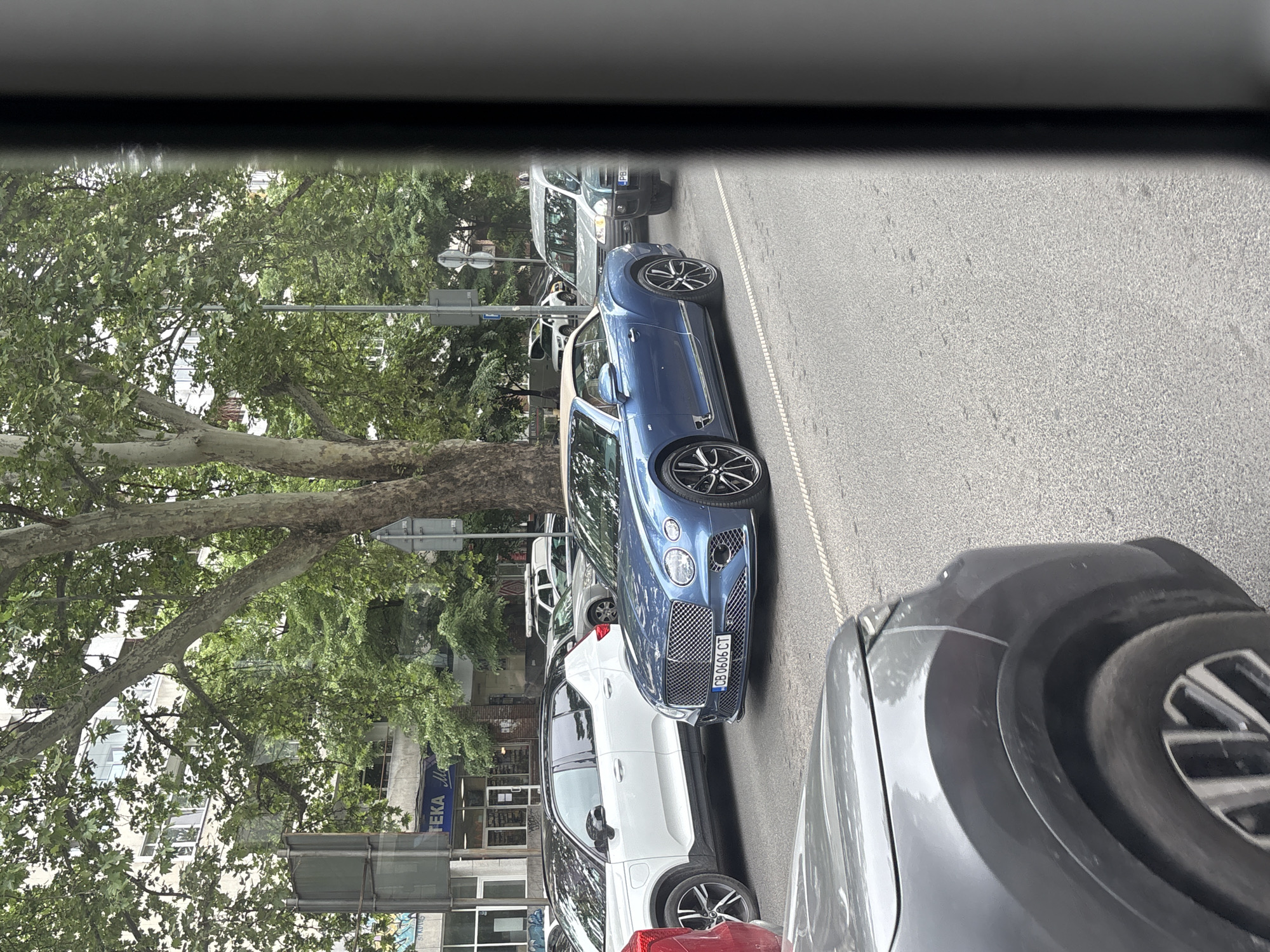 The image depicts a street scene in Plovdiv, Bulgaria. A blue Bentley convertible with license plate CB 0606 CT is parked on the side of the road. A white Volvo SUV is parked behind it. A gray car is visible in the foreground, obstructing part of the view. A building with a pharmacy (TEKA M) is visible in the background, along with trees and street signs. The photo appears to be taken from inside another vehicle.