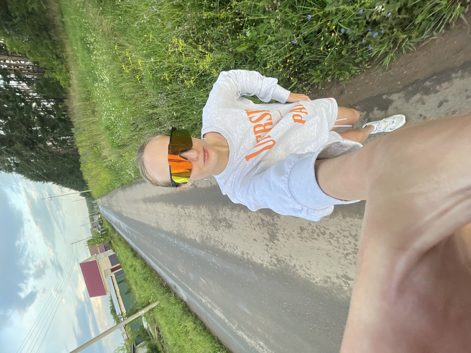 A young woman, wearing a grey sweatshirt and white shorts, with bright orange mirrored sunglasses, takes a selfie while walking on a wet, asphalt road. The road is lined with tall green grass and wildflowers on the right side and appears to lead into a village or rural area with houses and power lines visible on the left. The sky is cloudy, suggesting a potentially overcast or post-rain condition. The woman is in the foreground, her arm extending out to capture the shot, with the rest of her body and the environment stretching out behind her.