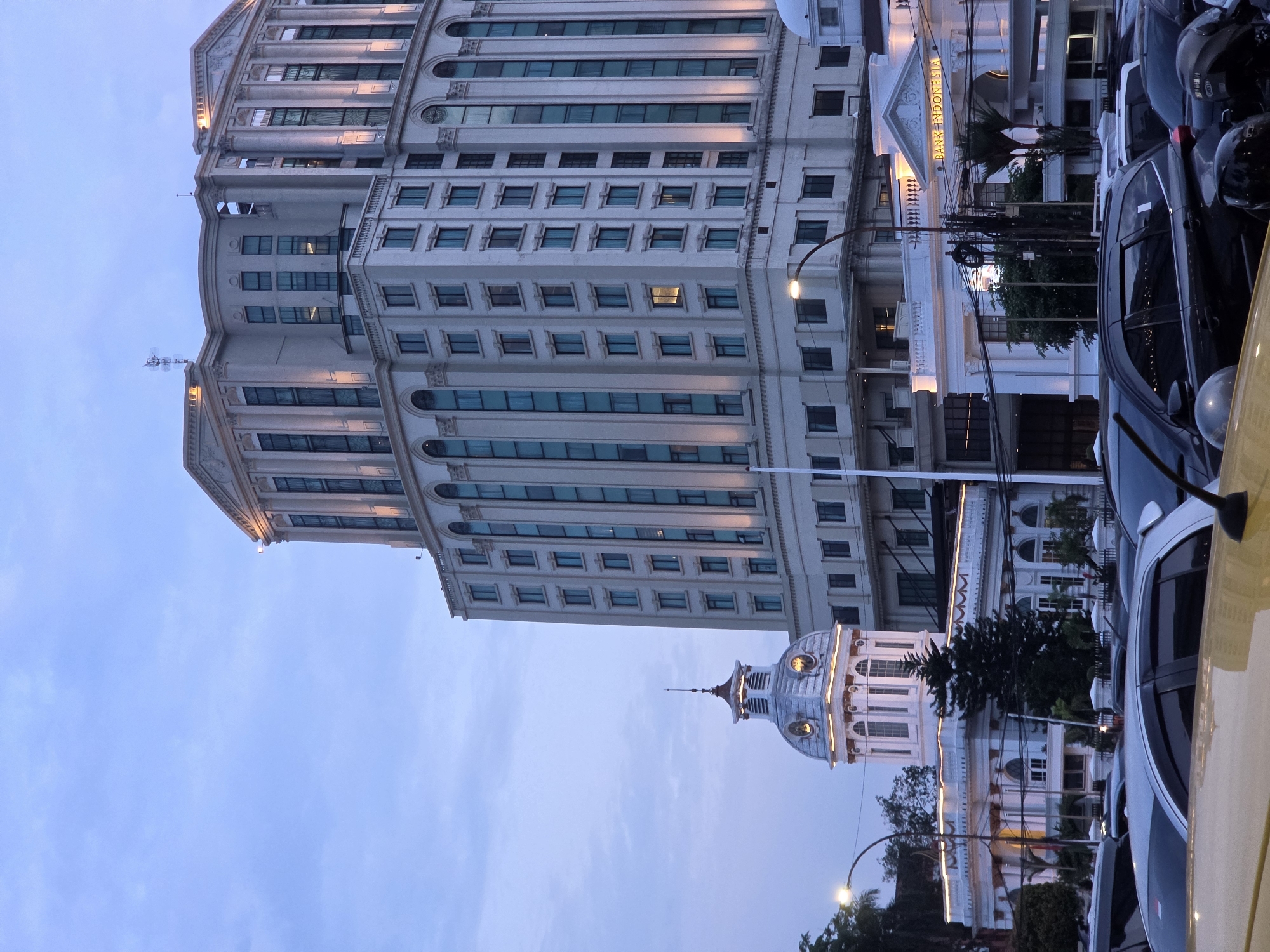 The image depicts a street scene in the City of Medan, Indonesia, at dusk. In the background, a tall, modern building with numerous windows and architectural embellishments dominates the skyline. To the left of this building is a smaller, white, classical-style structure with a prominent dome and clock tower. The facade of this building is illuminated, and it has the words "BANK INDONESIA" written above an entrance. In the foreground, a line of parked cars is visible, with the hoods of several vehicles taking up the bottom of the frame. The sky is a muted blue, indicating the transition from day to night. There are no people clearly visible in the image. The lighting suggests that the sun has recently set or is about to rise.