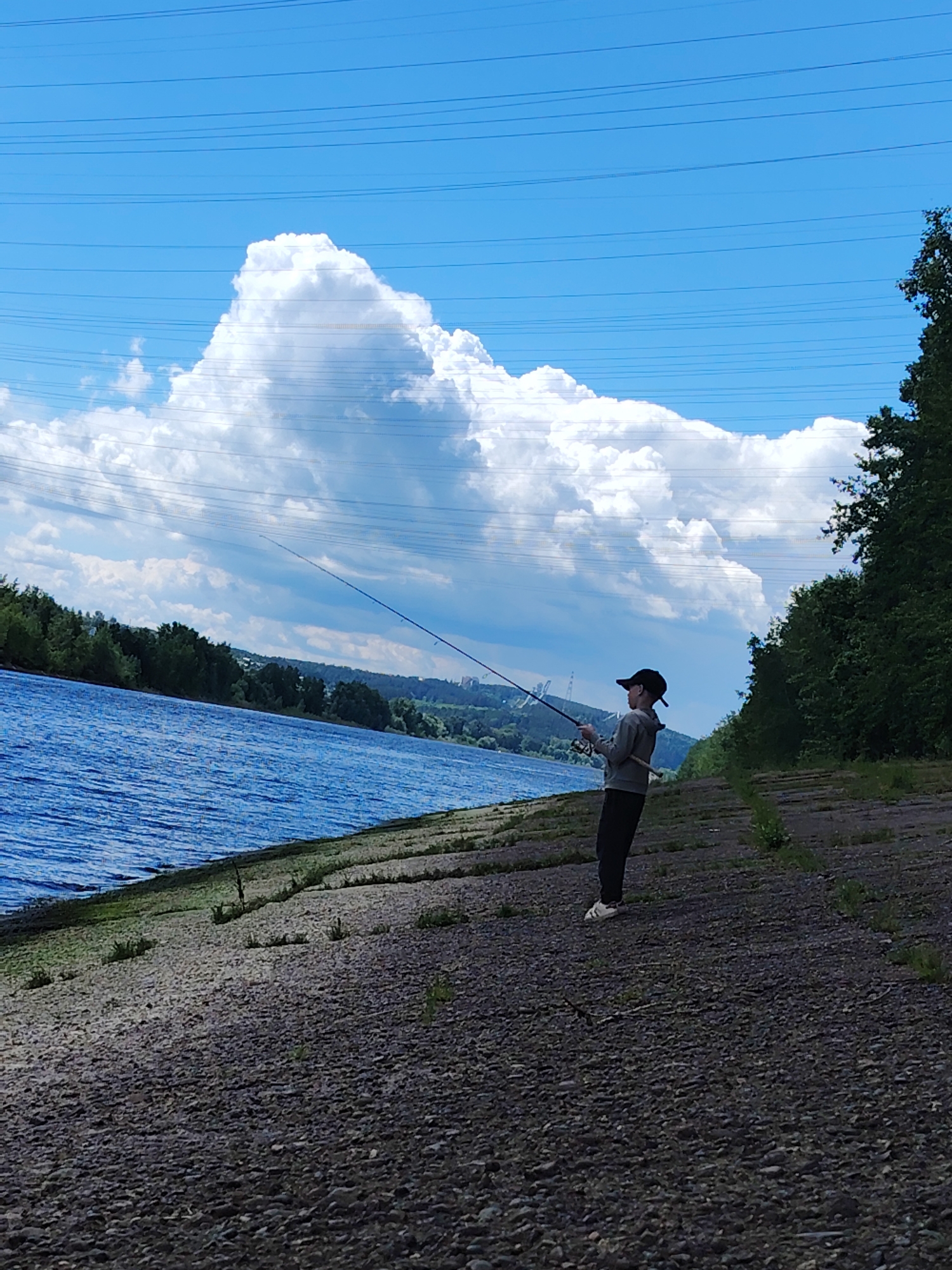 A person is fishing by a river in Chaikovsky, Russia on a clear, sunny day. The person is wearing a black baseball cap, a grey hooded sweatshirt, and dark pants. They are standing on a rocky embankment next to the blue water of the river, holding a fishing rod. The sky is blue with large, fluffy white clouds. Overhead power lines are visible against the sky. Lush green trees and vegetation line the riverbanks in the background, and a distant cityscape is barely visible on the horizon.
