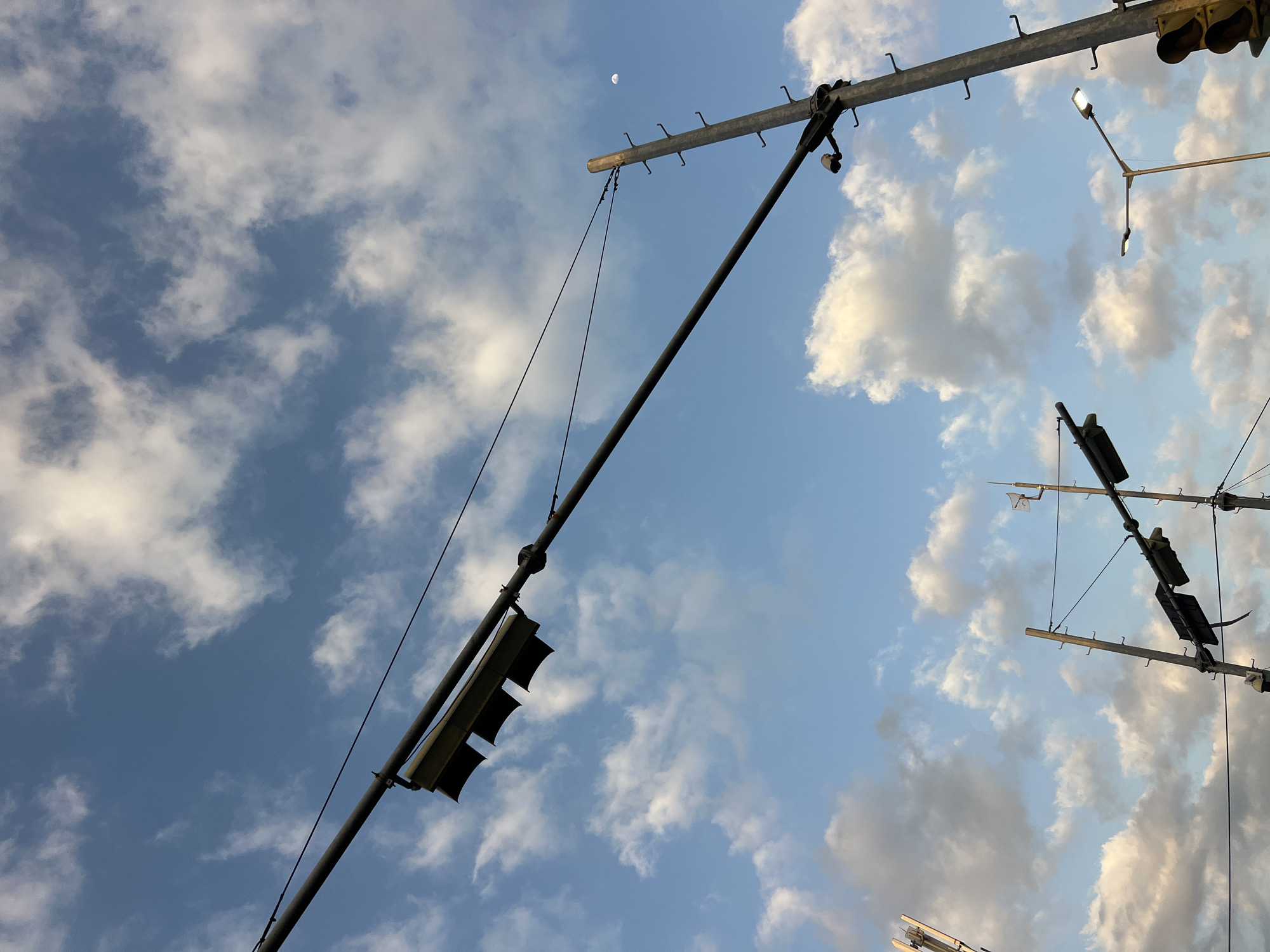 This is a low-angle, outdoor photograph taken on a partly cloudy day, looking up at a wide expanse of blue sky dotted with white cumulus clouds. In the foreground, a network of dark metal poles and overhead wires stretches across the frame. Several traffic signals are visible, mounted on the poles and suspended from the wires. Some of these signals are black and appear to be turned off, while one in the lower right corner shows a lit yellow signal. A small, crescent moon is faintly visible in the upper right quadrant of the sky. The scene appears to be an urban intersection, potentially in Denpasar Barat, Indonesia, given the context. No people are present in the image. The overall impression is one of urban infrastructure set against a natural sky.