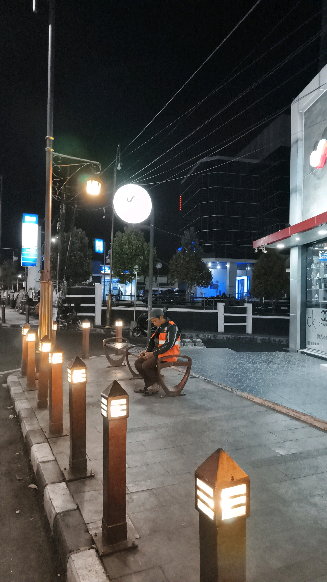 A person sits on a bench on a sidewalk at night. The person is wearing a grey baseball cap and an orange reflective vest over a dark shirt and pants. They are looking down at their hands. The bench is made of brown metal with a curved design. To the left of the bench are several square, brown bollard lights that are illuminated. The sidewalk is paved with gray tiles. In the background, across the street, is a building with a red awning and glass windows displaying merchandise. Further back, a large dark building with many illuminated windows rises into the night sky, crisscrossed by overhead wires. Trees are visible in the mid-ground. The overall scene suggests an urban environment in Boyolali, Indonesia, possibly a commercial or pedestrian area after closing hours.