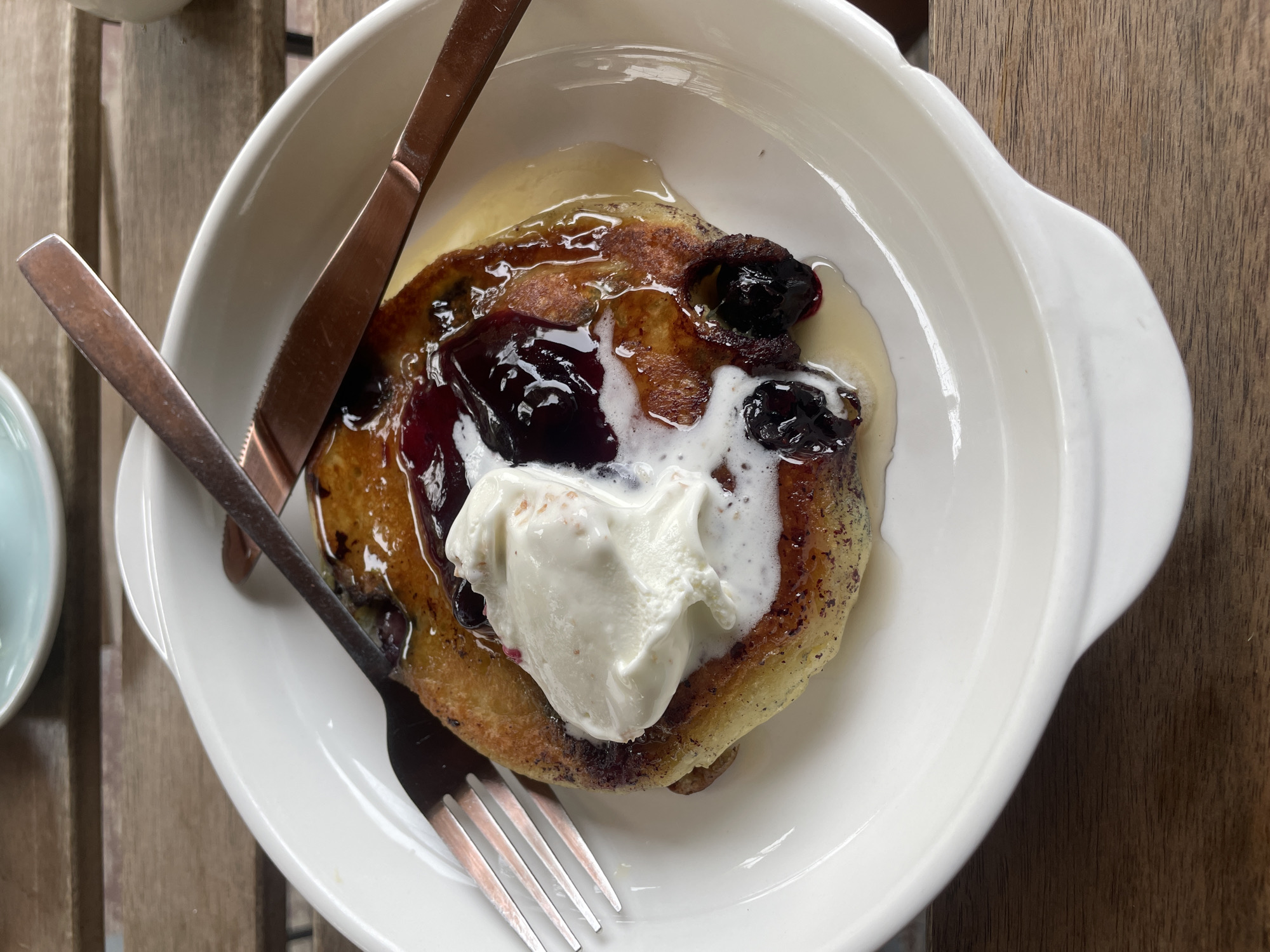 An overhead shot captures a single serving of a dessert or breakfast dish presented in a white ceramic bowl with small handles, resting on a slatted wooden table. The central subject is a golden-brown, thick pancake or French toast-like item, generously topped with a dark berry compote or fresh berries, possibly blueberries or a mix of dark fruits. A dollop of white, soft cream, likely whipped cream or ice cream, sits on top of the berries, slightly melting into the warm dish. A light, creamy sauce, possibly custard or vanilla syrup, pools around the base of the dish within the bowl.
A knife and fork with distinctive rose gold or copper-colored handles are placed within the bowl, indicating the dish is ready to be eaten. The wooden table beneath has a natural grain and distinct planks, suggesting a rustic or outdoor cafe setting. The lighting is bright and natural, casting soft shadows, consistent with daylight. The overall presentation is appealing and suggests a delightful culinary experience in Chiang Mai City Municipality, Thailand. There is no visible text in the image.