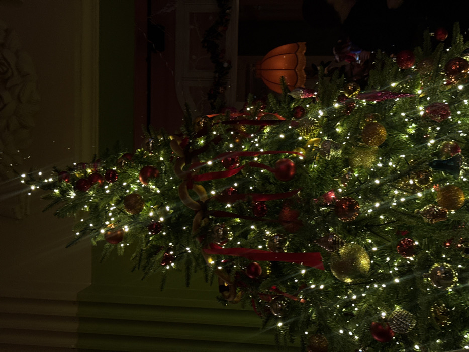 The image displays a large, elaborately decorated Christmas tree illuminated by numerous small white lights, positioned indoors within a dimly lit, formal room in Yekaterinburg, Russia. The tree is densely adorned with a variety of ornaments, predominantly spherical in red, gold, and silver tones, along with draped red ribbons and gold bows.
To the left of the tree, a dark green curtain or drapery extends vertically, meeting a lighter-colored ceiling featuring ornate, possibly molded, architectural details. To the right, a section of a dark red wall is visible, below which a white or light-colored fireplace mantelpiece is discernible, decorated with a dark garland. An orange-tinted, bell-shaped lampshade hangs in the mid-ground, to the right of the tree. A faint, dark silhouette of a person, showing a head and shoulders, is partially visible in the lower right quadrant, behind the tree. The environment suggests an upscale interior, consistent with a grand hotel, museum, or historic private residence. No specific actions or interactions are clearly visible beyond the static scene.