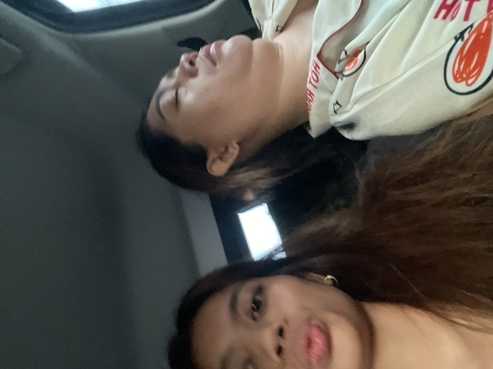 The image depicts two women inside a vehicle, likely in motion given the blurred background. One woman is facing the camera with brown hair, while the other is lying down, possibly asleep, wearing a white pajama-style top with red accents. The vehicle has a sunroof visible at the top of the frame. There are no immediate indicators of the location being Davao City, Philippines.